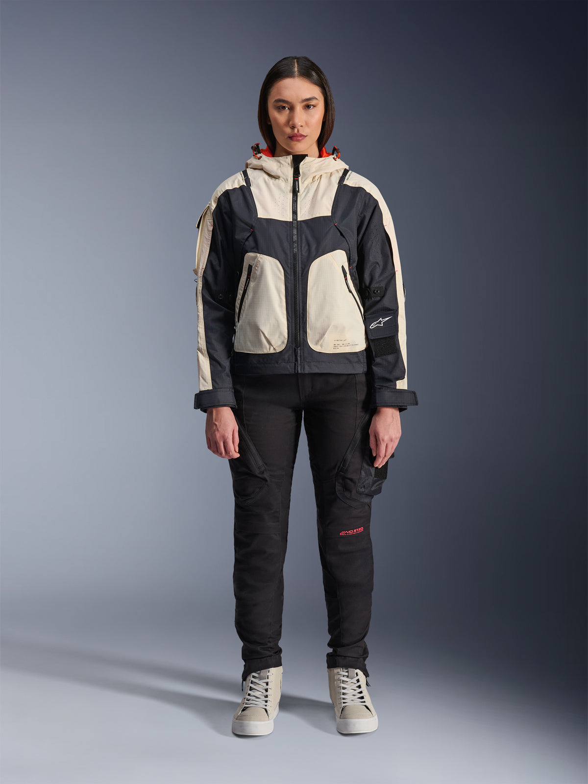 Modella che indossa la giacca Alpinestars Molly Air in Black Almond, con design color-block crema e nero e cappuccio foderato in rosso, abbinata a pantaloni da moto neri e sneakers crema, scatto in studio di abbigliamento urban contemporaneo.