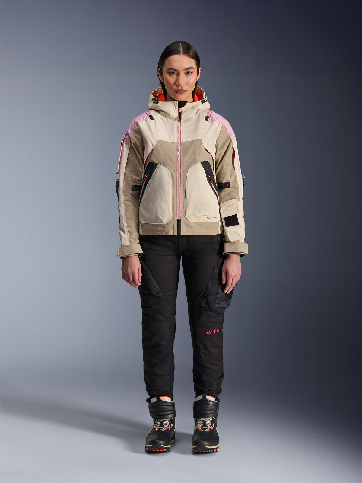 Modella che indossa la giacca Alpinestars Molly Air in Sand Pink Almond, abbinata a pantaloni tecnici da moto e stivali neri, vista frontale a figura intera, giacca da moto urbana con design d'ispirazione utility e accenti rosa