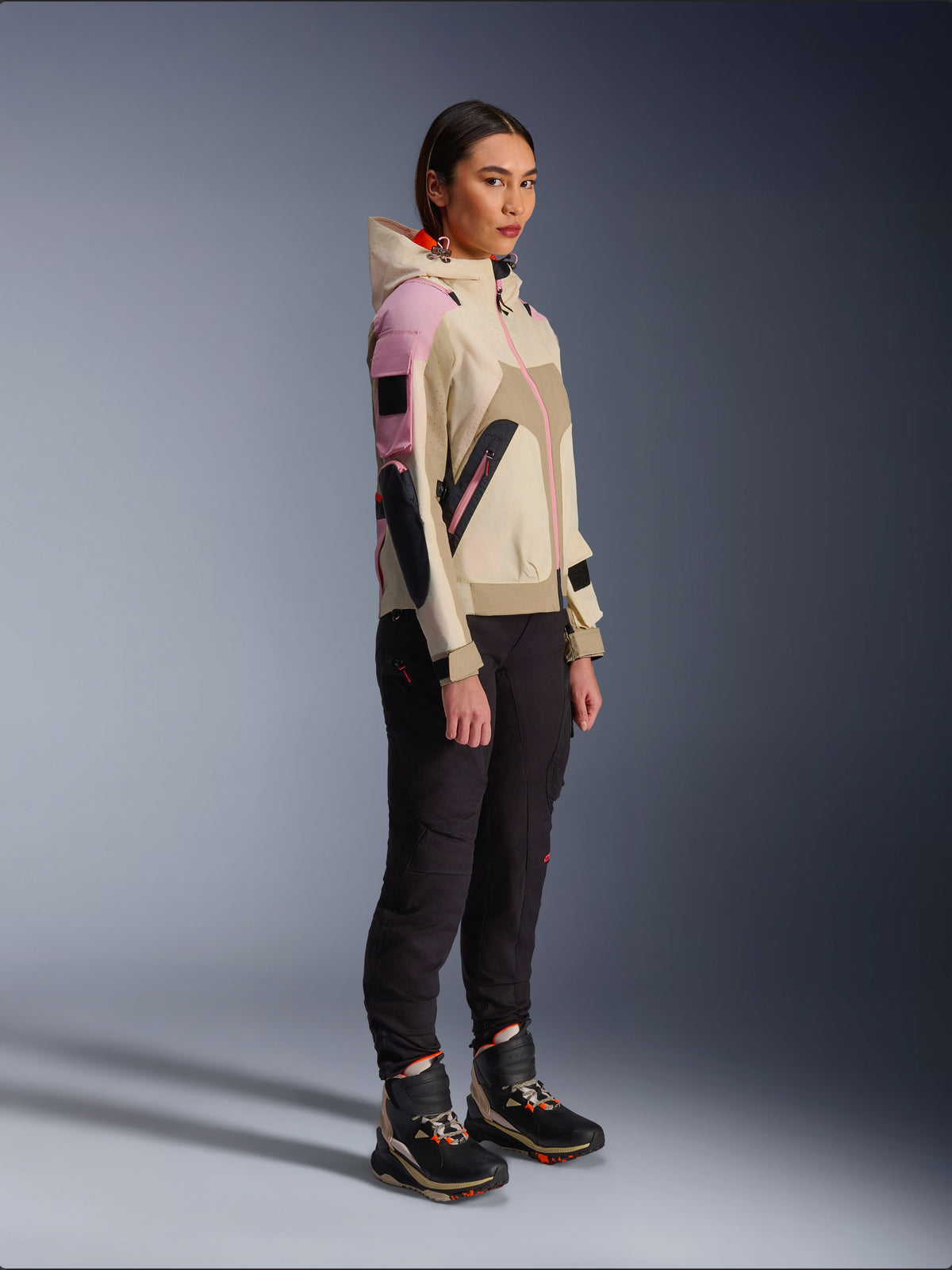 Modella che indossa la giacca Alpinestars Molly Air in Sand Pink Almond, abbinata a pantaloni tecnici scuri e stivali da moto alti, giacca da moto urbana con cappuccio e pannelli multicolore, design contemporaneo ispirato allo streetwear