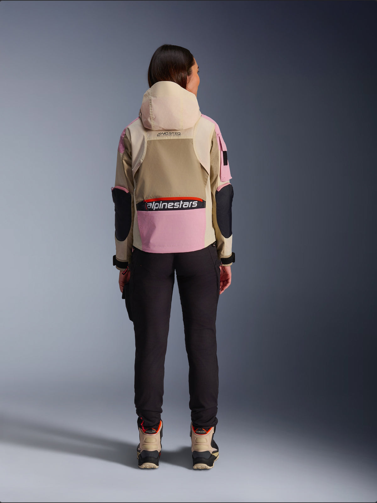 Motociclista vista da dietro che indossa la giacca Alpinestars Molly Air in Sand Pink Almond, con cappuccio beige e pannelli rosa con dettagli neri, abbinata a pantaloni da moto neri e stivali beige, abbigliamento da moto urban contemporaneo