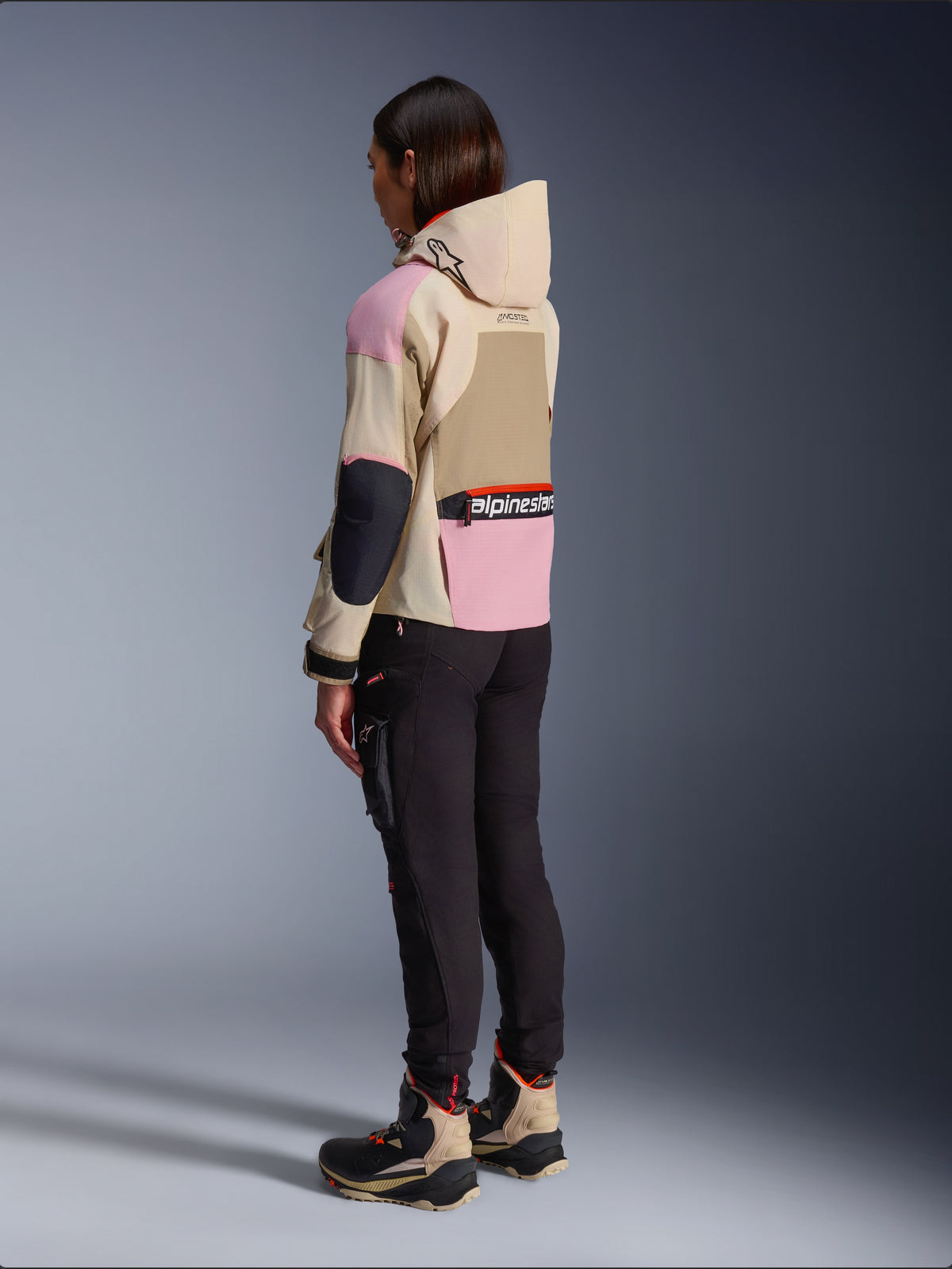 Motociclista che indossa la giacca Alpinestars Molly Air in Sand Pink Almond, caratterizzata da un design multi-tonale beige e rosa, mostrata da una vista laterale posteriore con pantaloni da moto neri e stivali tecnici abbinati, stile motociclistico urbano con cappuccio.