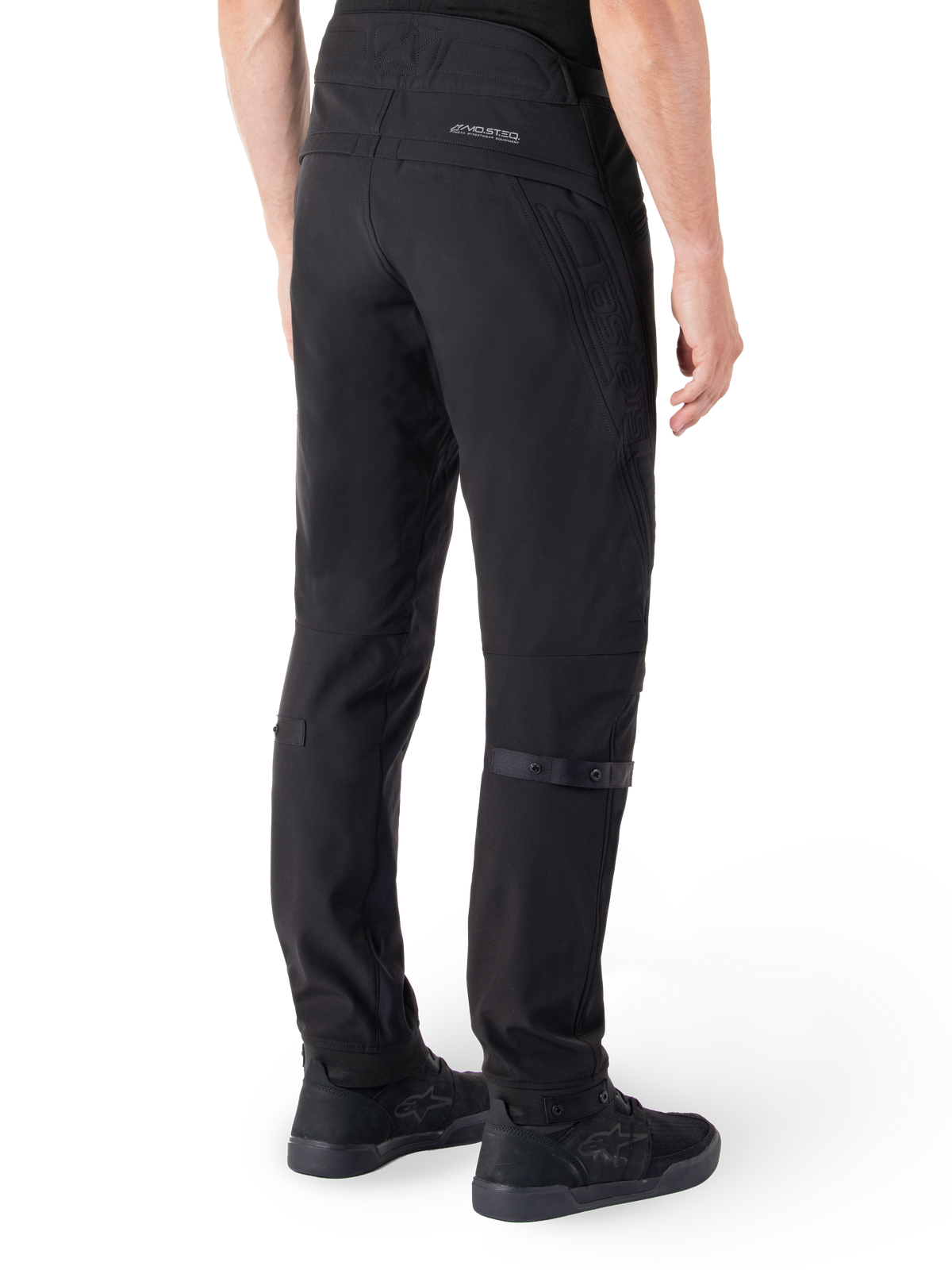 Alpinestars Alden Pants, pantaloni da moto urban, nero, vista posteriore che mostra il tessuto softshell tecnico con dettagli trapuntati, branding riflettente MO.ST.EQ. sul carré posteriore e cinturini regolabili sui polpacci, abbinati a scarpe da guida nere