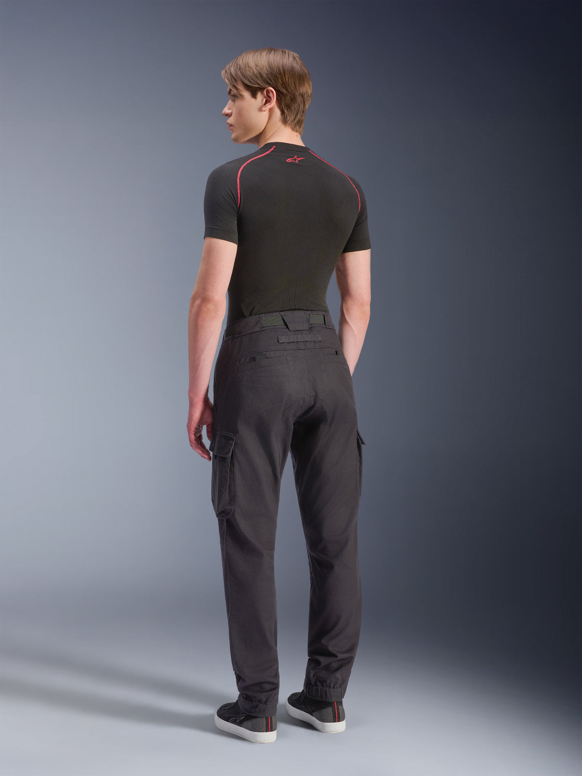 Alpinestars Flex-AST Cargo Pants - Short, pantaloni cargo da moto, nero, vista posteriore di un modello che indossa pantaloni cargo con vestibilità regolare affusolata, tasche laterali e orli elasticizzati, abbinati a un base layer nero e scarpe casual