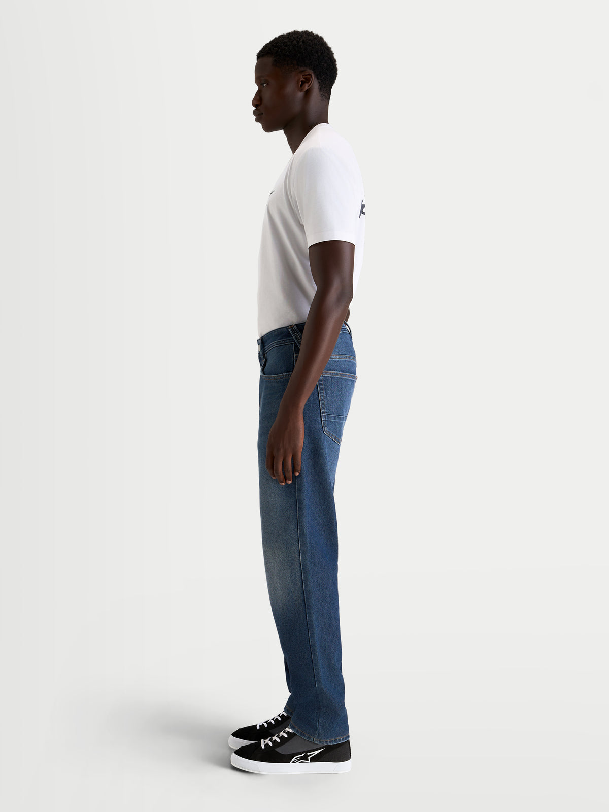 Alpinestars Flex Ast 8 Denim Pantaloni, pantaloni da moto urban da uomo in blu medio, vista laterale su un modello con maglietta bianca e scarpe casual da moto, denim monostrato elasticizzato di classe AA