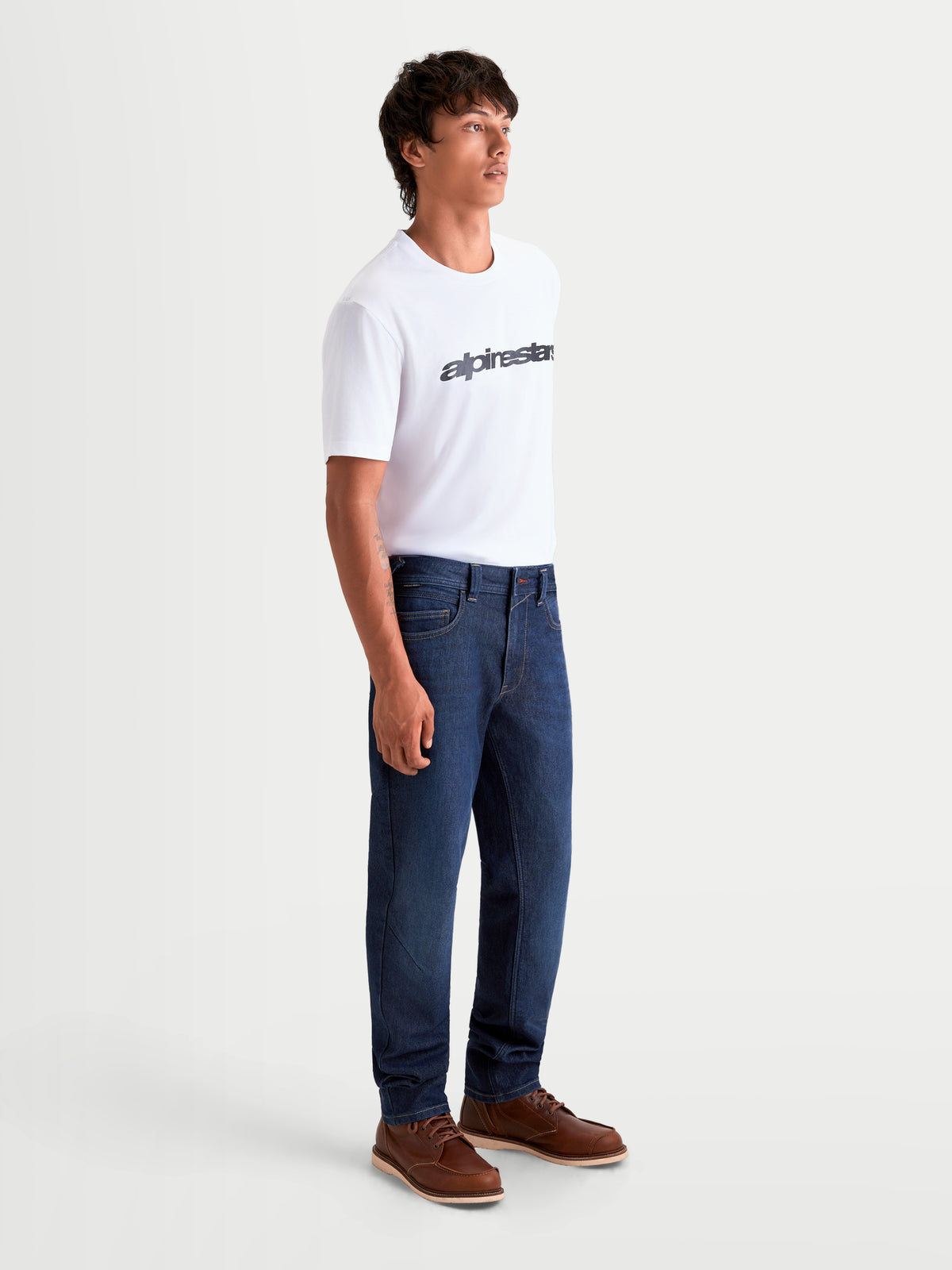 Alpinestars Flex Ast 7 Denim Pantaloni, pantaloni da moto urbani contemporanei da uomo in blu scuro, vista laterale su un modello che indossa una t-shirt bianca e stivali marroni, abbigliamento da moto monostrato certificato AA con comfort elasticizzato
