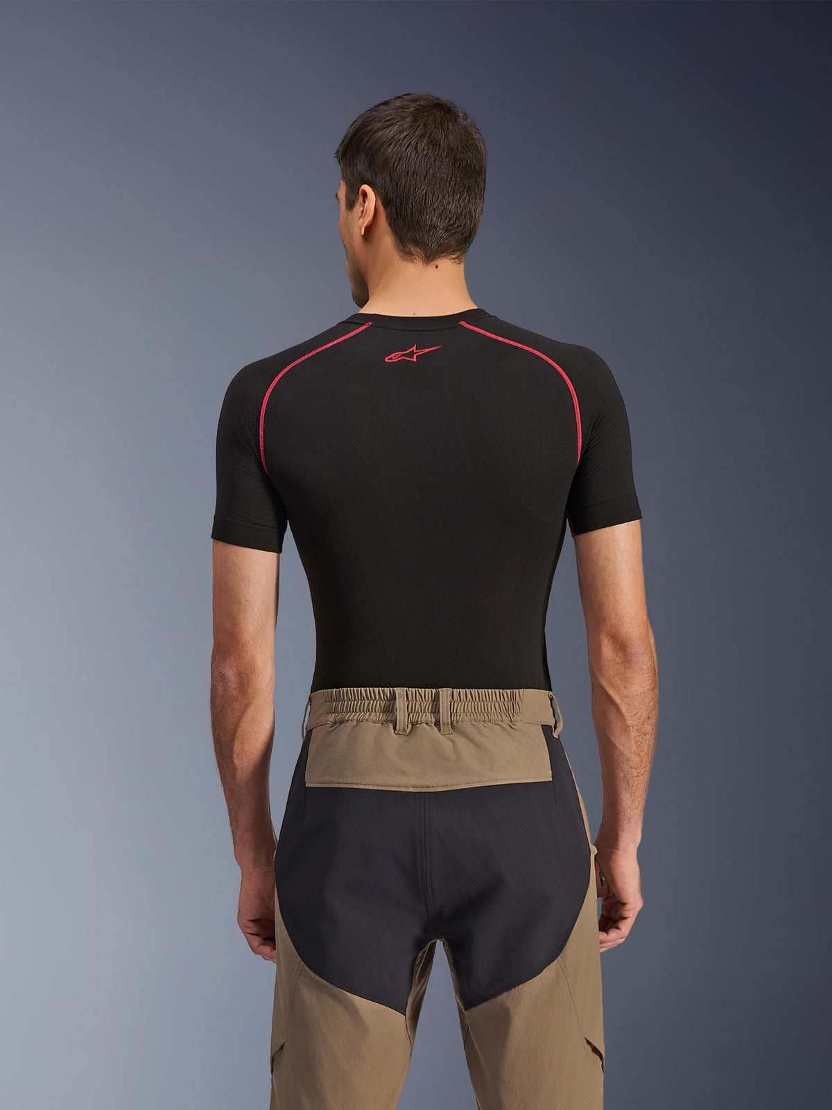 Alpinestars Flex-AST Explorer Pants, vista posteriore su modello, colore Walnut Black, caratterizzati da tessuto marrone con pannelli rinforzati neri sulla seduta e vita elasticizzata, abbinati a un base layer nero a maniche corte con cuciture rosse.