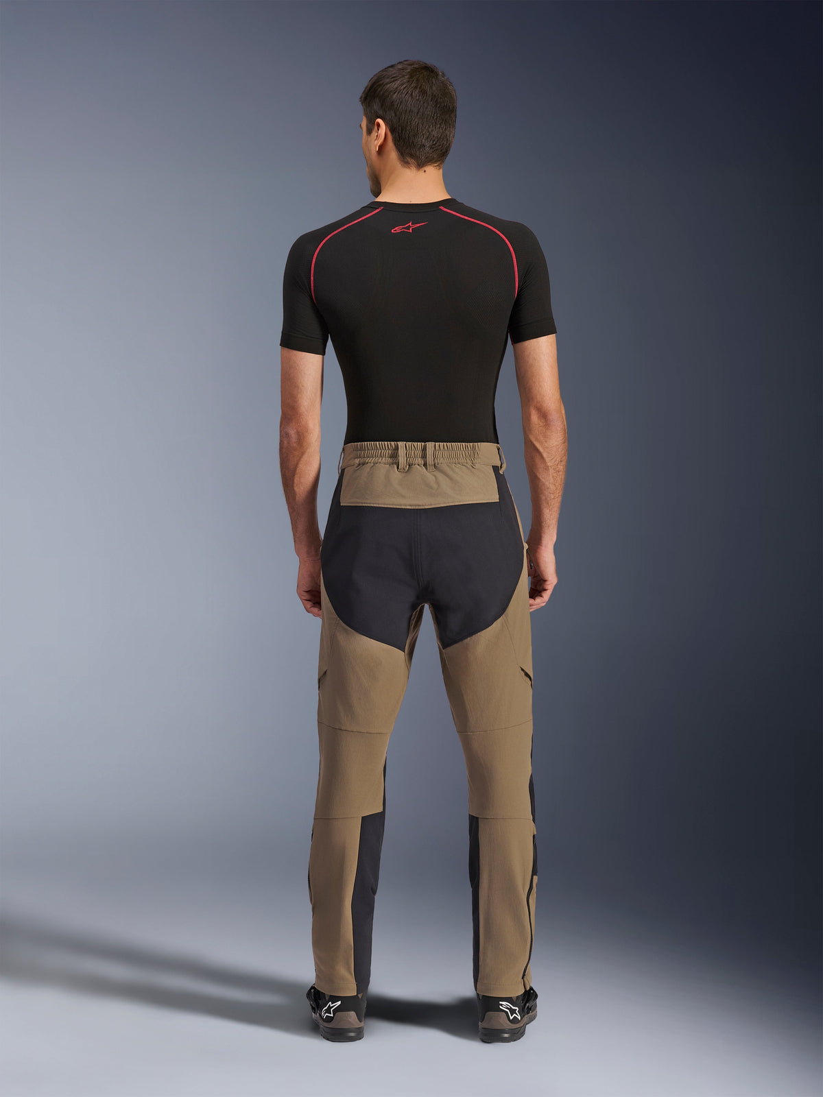 Pantaloni Alpinestars Flex-AST Explorer, Walnut Black, vista posteriore su modello, con seduta nera rinforzata e tessuto elasticizzato marrone, abbinati a base layer nero e stivali tecnici da moto, abbigliamento resistente per adventure riding