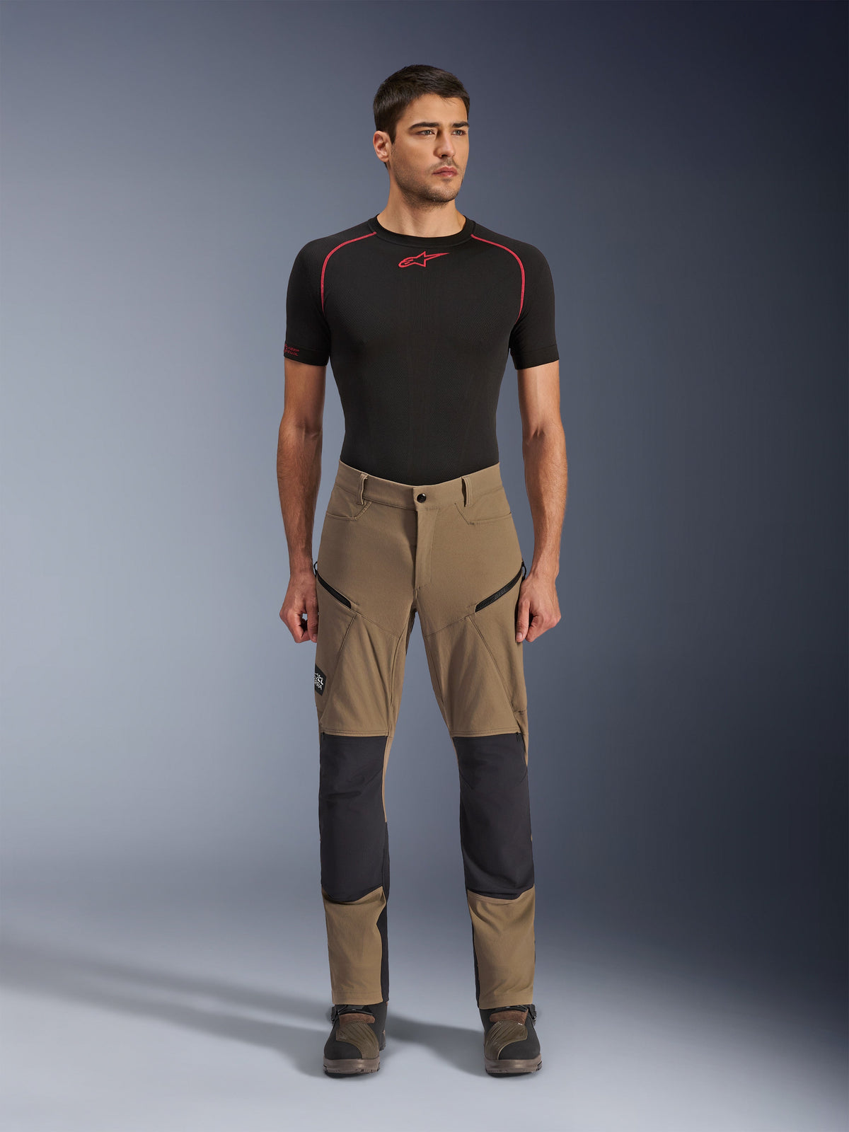 Pantaloni Alpinestars Flex-AST Explorer, pantaloni da moto touring, marrone walnut e nero, con pannelli rinforzati sulle ginocchia e tasche con zip sulle cosce, indossati con base layer nero a maniche corte, vista a figura intera