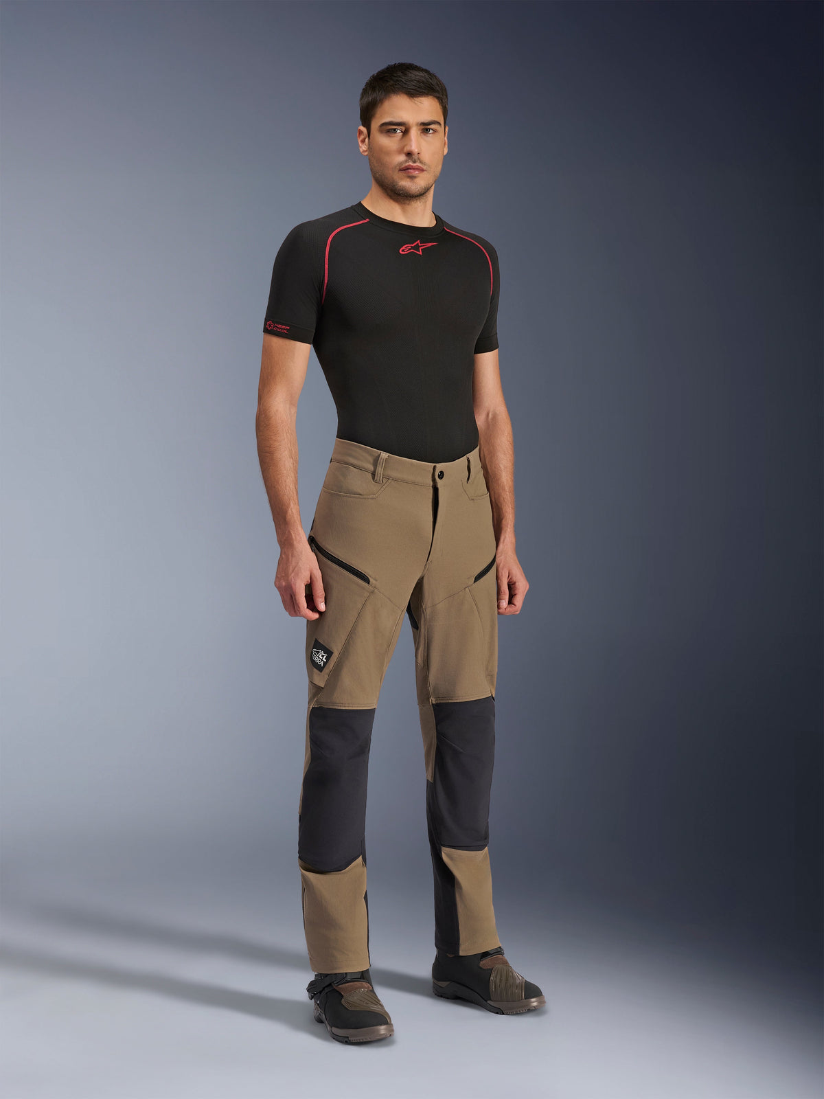 Pantaloni Alpinestars Flex-AST Explorer, pantaloni da moto adventure Walnut Black, indossati da un modello uomo con base layer nero a maniche corte e stivali adventure, con tasche cargo con zip e pannelli rinforzati sulle ginocchia, abbigliamento tecnico per piloti dual-sport e touring