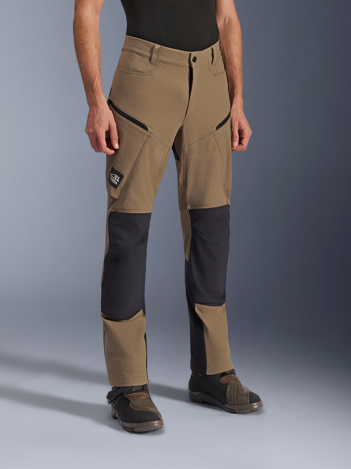 Alpinestars Flex-AST Explorer Pants, pantaloni da moto adventure in Walnut Black, in tessuto color sabbia con pannelli neri rinforzati su ginocchia e seduta, tasche cargo con cerniera e patch logo All Terra, mostrati indossati con stivali tecnici da guida.