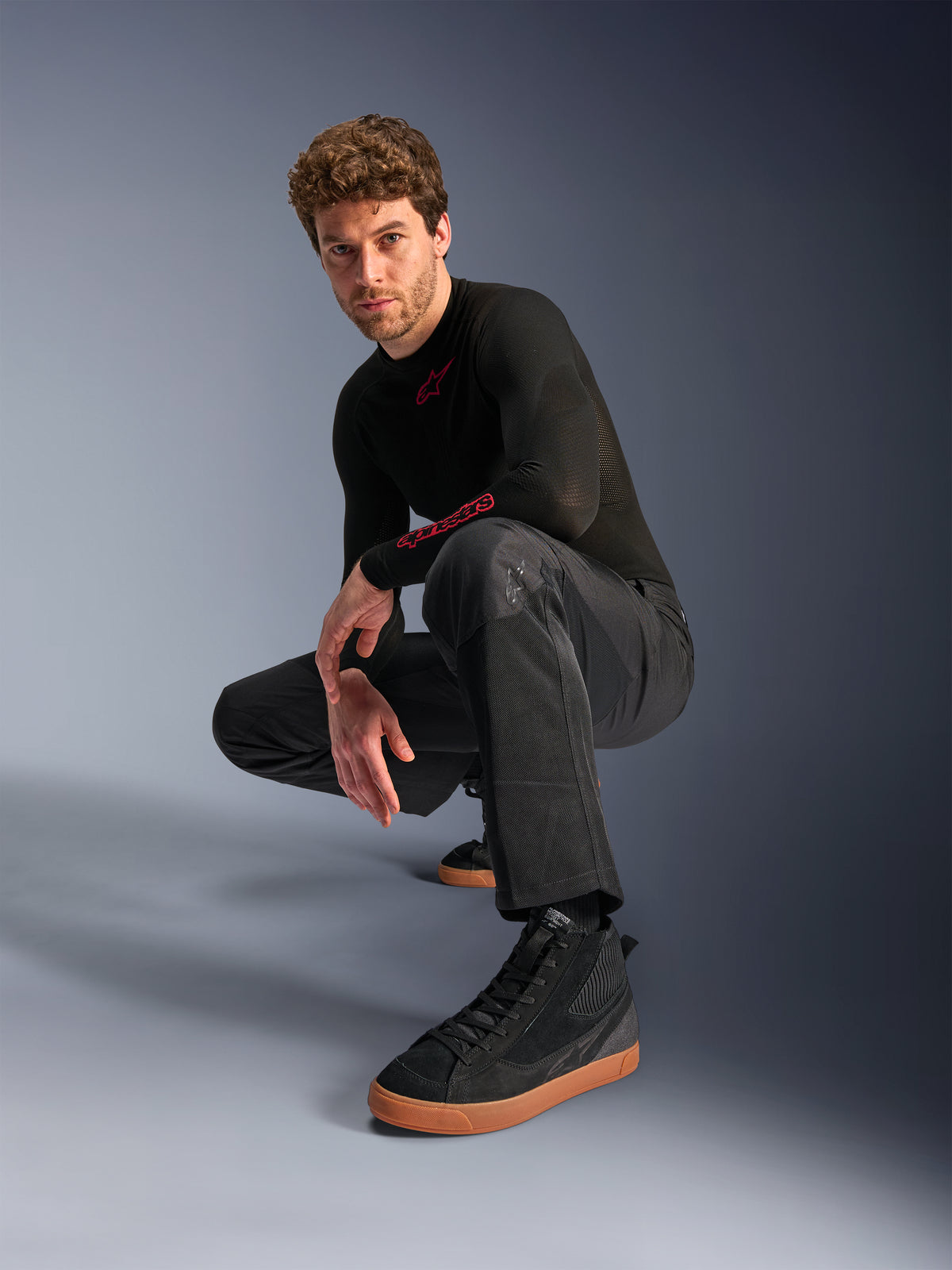 Alpinestars C-1 Air Pants, pantaloni da moto urban, nero, modello accovacciato in studio, indossati con maglia nera a maniche lunghe e sneakers nere con suole marroni, dotati di pannelli in mesh traspirante e stile urban contemporaneo