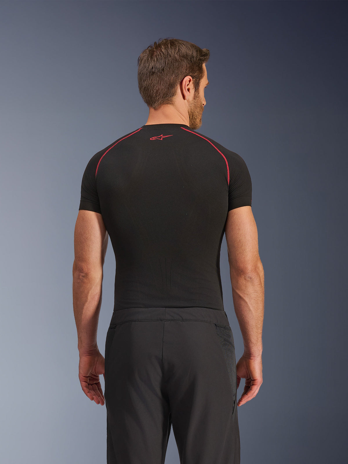 Vista posteriore di un modello che indossa i pantaloni Alpinestars Aeroshell Airflow, neri, costruzione leggera in softshell con pannelli laterali in rete integrati, abbinati a un base layer nero con cuciture rosse a contrasto, abbigliamento sportivo da moto