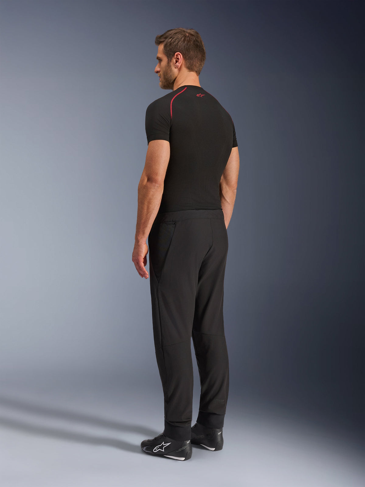 Pantaloni Alpinestars Aeroshell Airflow, pantaloni da moto neri, leggero tessuto softshell, indossati con maglia tecnica nera a maniche corte e stivali da moto neri, vista laterale posteriore che mostra la vestibilità slim e la costruzione ergonomica