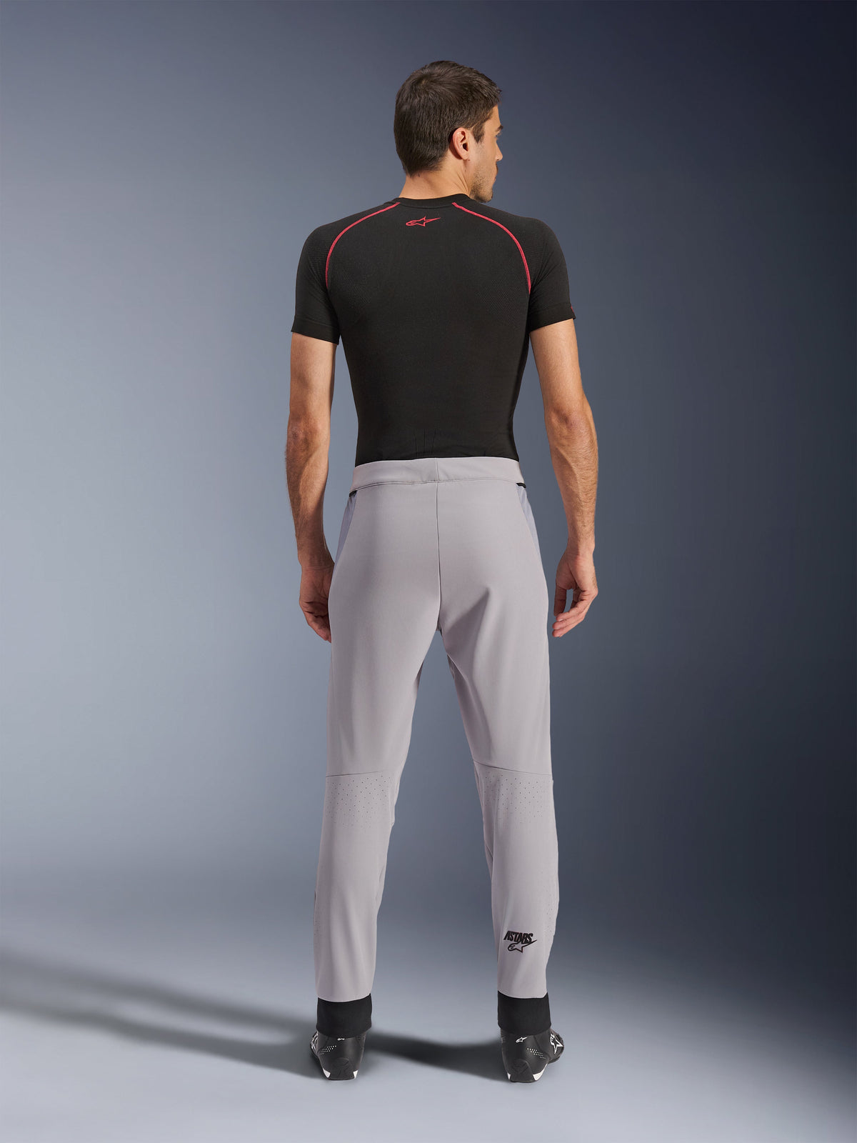 Pantaloni Alpinestars Aeroshell Airflow, Mid Gray, vista posteriore con zone perforate al laser sui polpacci, costruzione in softshell elasticizzato leggero, indossati con maglia nera a maniche corte e stivali da moto, design sportivo per climi caldi