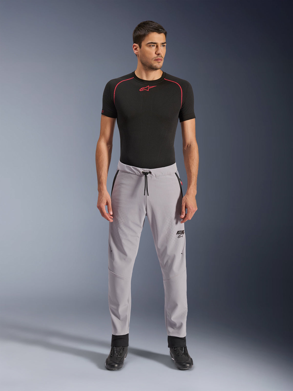 Alpinestars Aeroshell Airflow Pants, Grigio Medio, pantaloni in tessuto leggero con zone di ventilazione traforate al laser e vita con coulisse, indossati da un modello con maglia a compressione nera e stivali da moto neri, design sportivo moderno