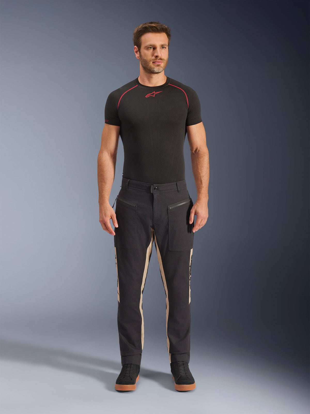 Modello uomo con pantaloni Alpinestars Flex-AST Canvas in Black Aluminum, pantaloni da moto urban, neri con pannelli beige, dotati di tasche utility e cerniere ispirate al workwear, abbinati a base layer nero a maniche corte e sneakers nere