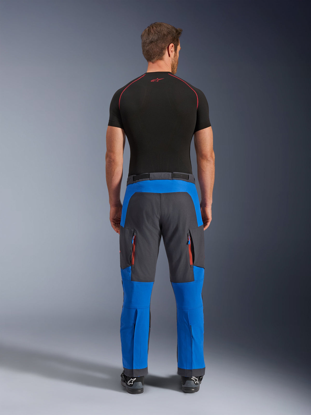 Pantaloni Alpinestars Maxdura Dual in Blu Royal e Grigio Scuro, pantaloni da moto adventure visti dal retro, con tasche cargo con cerniera e accenti arancioni, design sopra lo stivale, indossati con un base layer nero a maniche corte