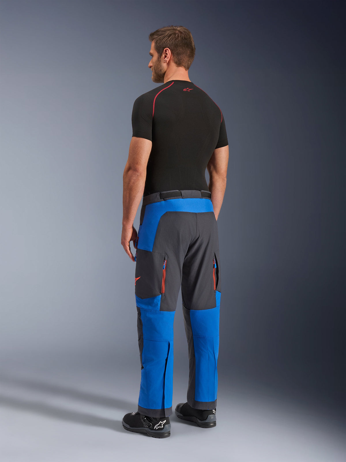 Pantaloni Alpinestars Maxdura Dual, pantaloni da moto adventure, blu reale e grigio scuro, vista posteriore indossata da un modello, con prese d'aria con zip arancioni, vestibilità adventure sopra lo stivale e materiale elasticizzato resistente.