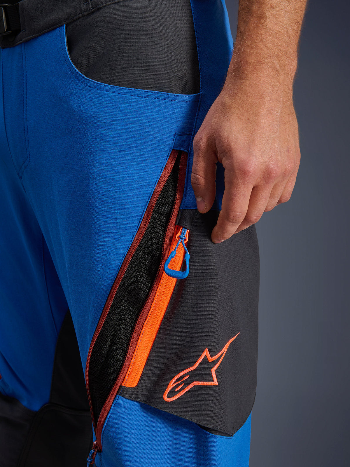 Dettaglio pantaloni Alpinestars Maxdura Dual, pantaloni da moto adventure blu royal e grigio scuro, primo piano della ventilazione con zip sulla coscia con accenti arancioni e linguetta blu, logo Alpinestars arancione su pannello nero, costruzione in poliammide elasticizzata resistente