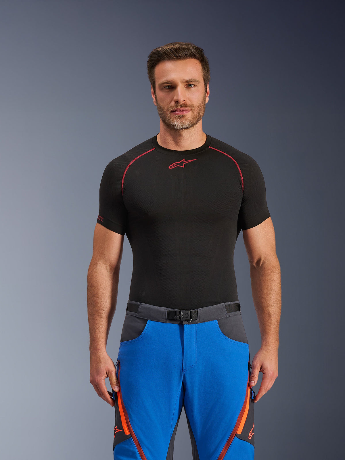 Pantaloni Alpinestars Maxdura Dual, blu royal e grigio scuro, pantaloni da moto adventure con dettagli arancioni, indossati da un modello con una maglia a compressione nera, dotati di cintura con fibbia magnetica e prese d'aria con zip sulle cosce, abbigliamento professionale per motorsport