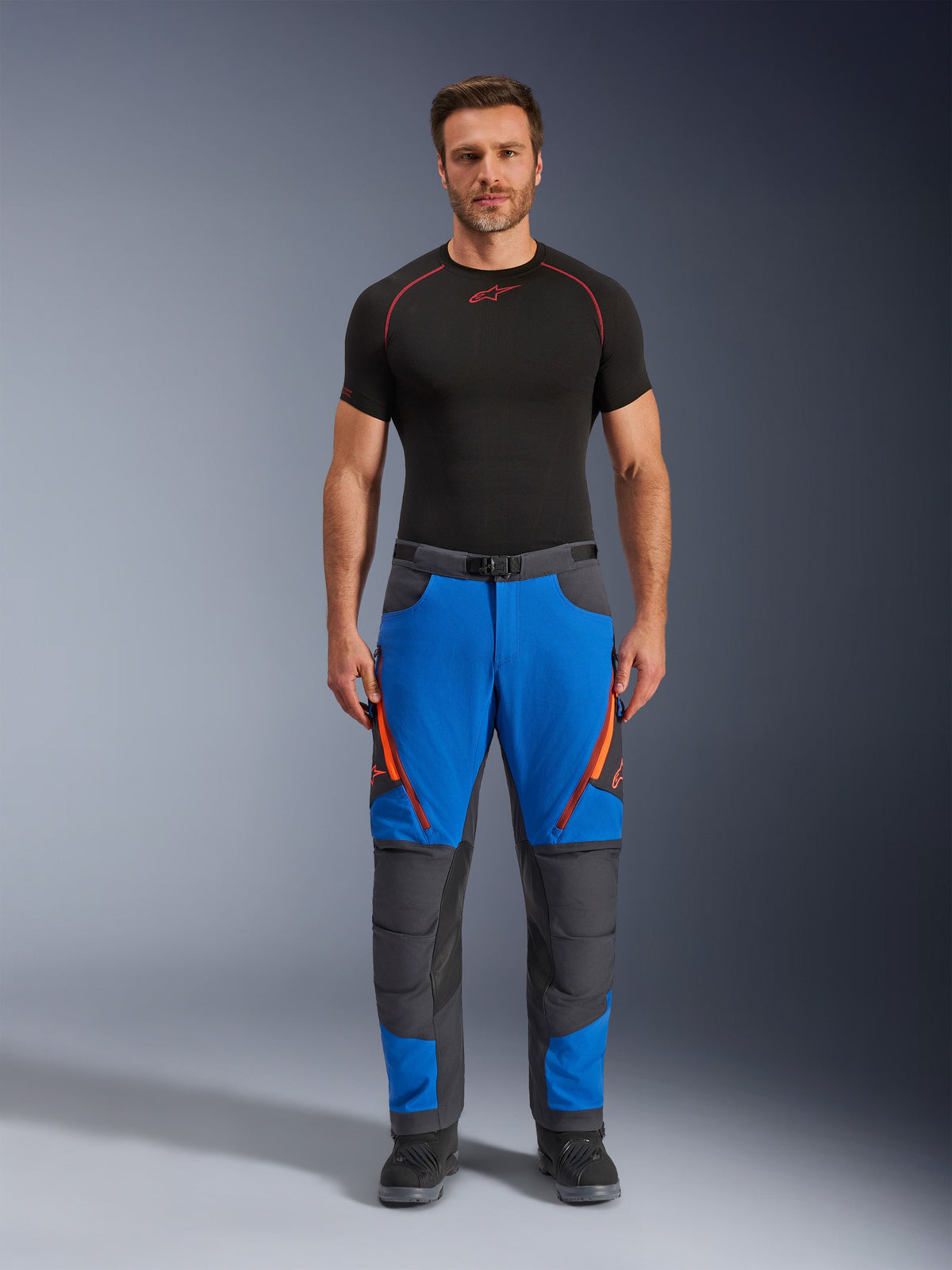 Alpinestars Maxdura Dual Pants, pantaloni da moto adventure, blu reale e grigio scuro, accenti con cerniere arancioni, indossati con base layer Alpinestars nero e stivali neri, con pannelli rinforzati sulle ginocchia e design tecnico da fuoristrada