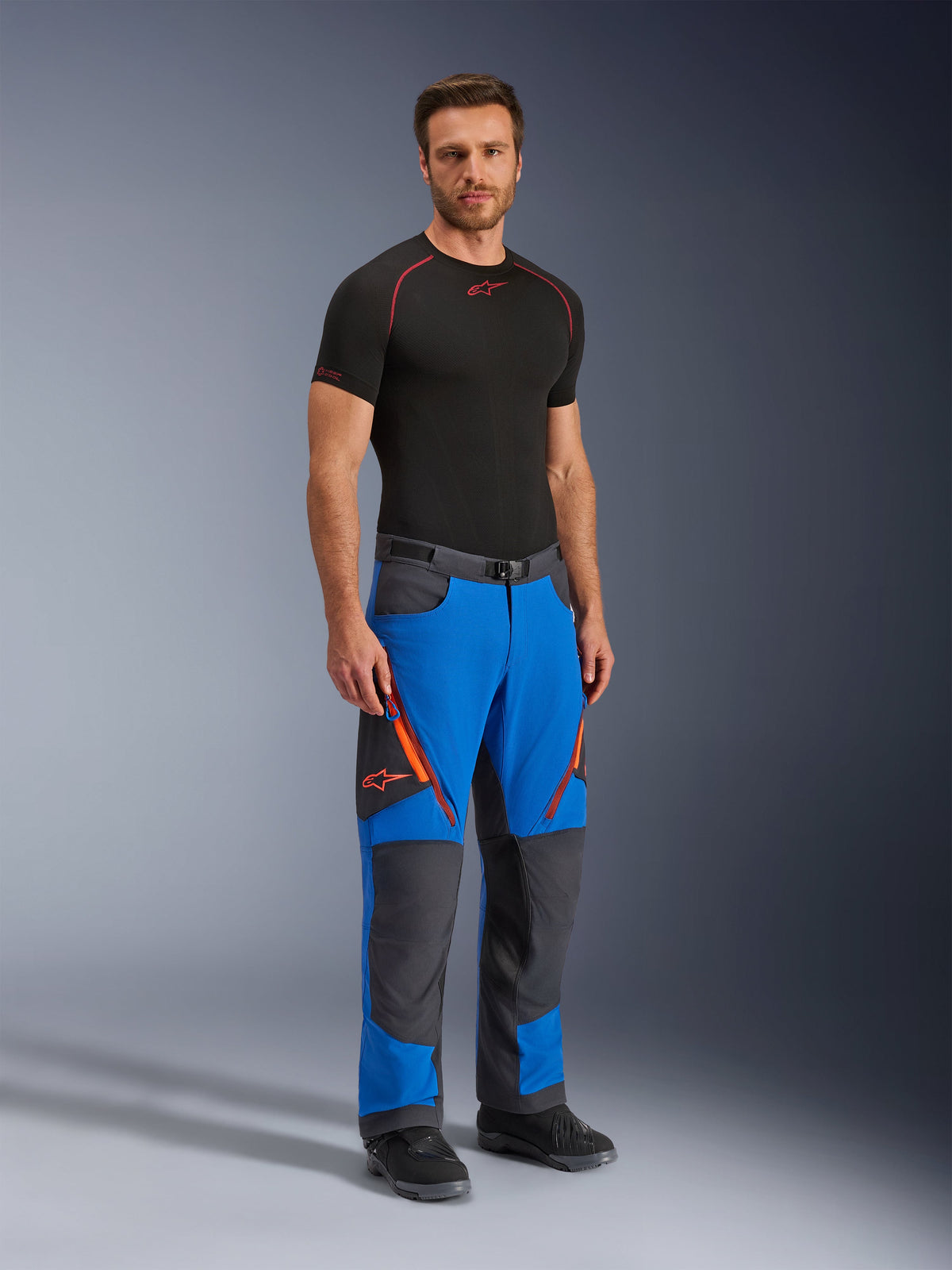 Alpinestars Maxdura Dual Pants, pantaloni da moto adventure, blu reale e grigio scuro, con dettagli delle cerniere e loghi arancioni, indossati da un modello con base layer e stivali neri, design tecnico con pannelli rinforzati per guida dual-sport