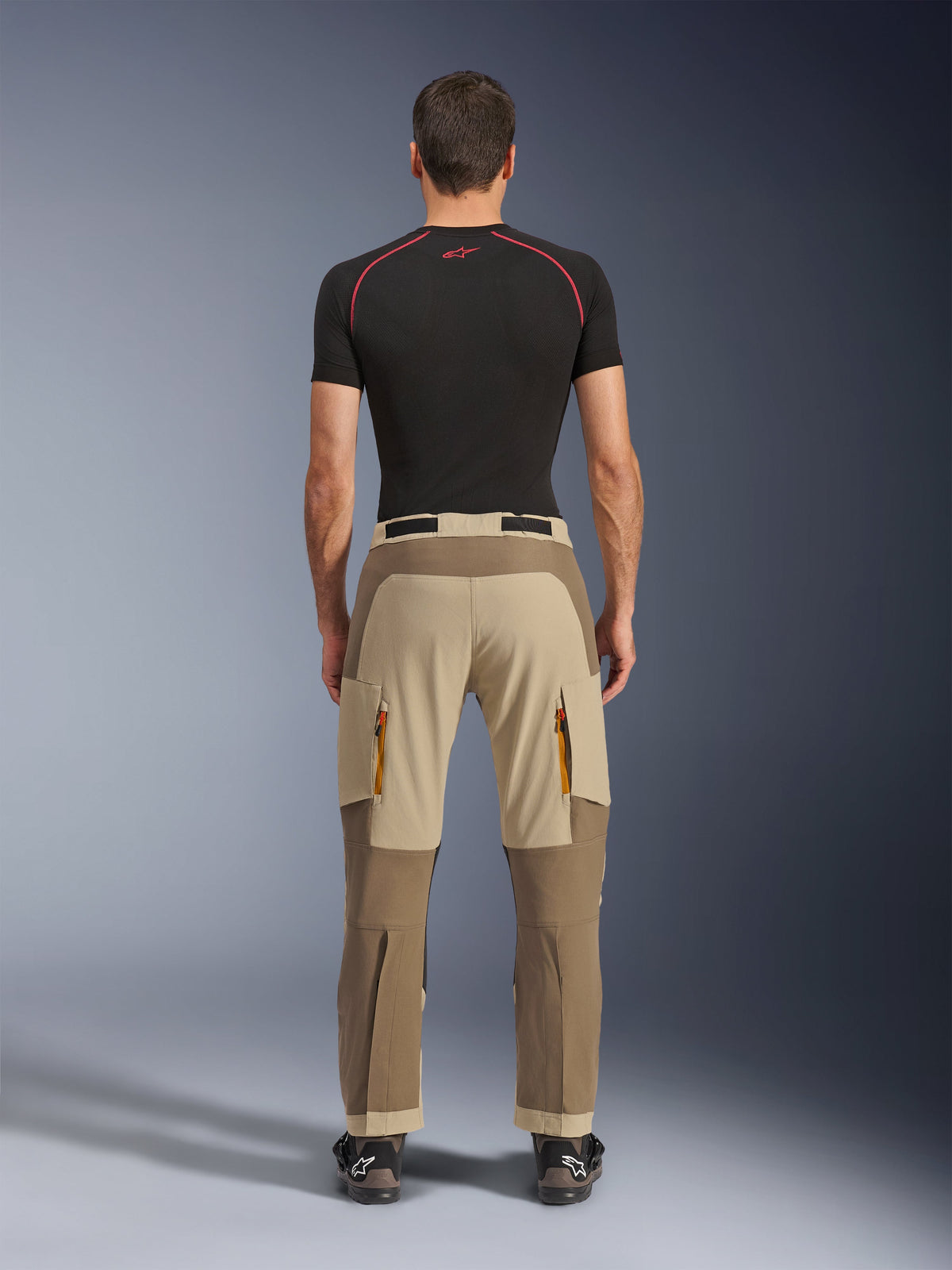 Alpinestars Maxdura Dual Pants in Walnut Aluminum, vista posteriore che mostra il design bicolore marrone e beige, pantaloni da moto adventure con tasche cargo con zip e seduta rinforzata, indossati con maglia a compressione nera e stivali da moto