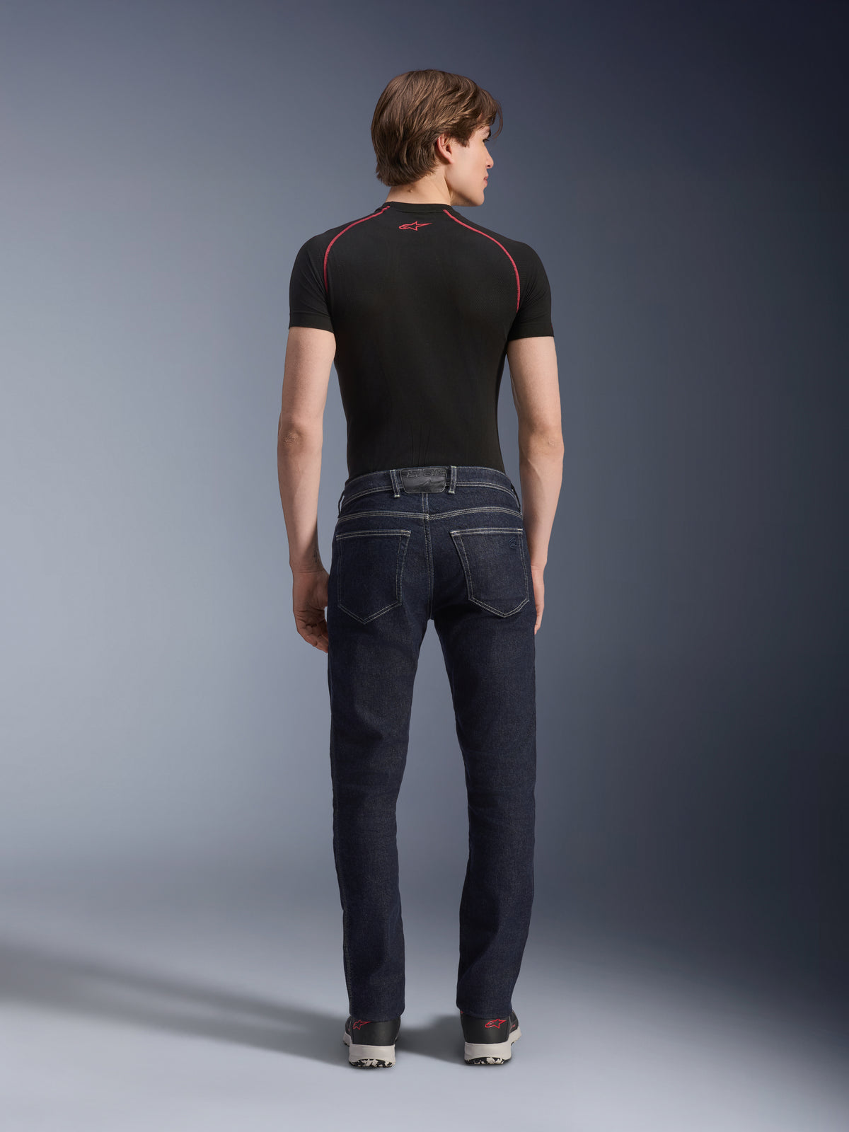 Pantaloni Alpinestars Copper V3 Denim in Rinse Blue, jeans da moto urban visti dal retro, caratterizzati da una vestibilità regular a gamba dritta e tasche posteriori, indossati con un top nero a maniche corte e sneakers.