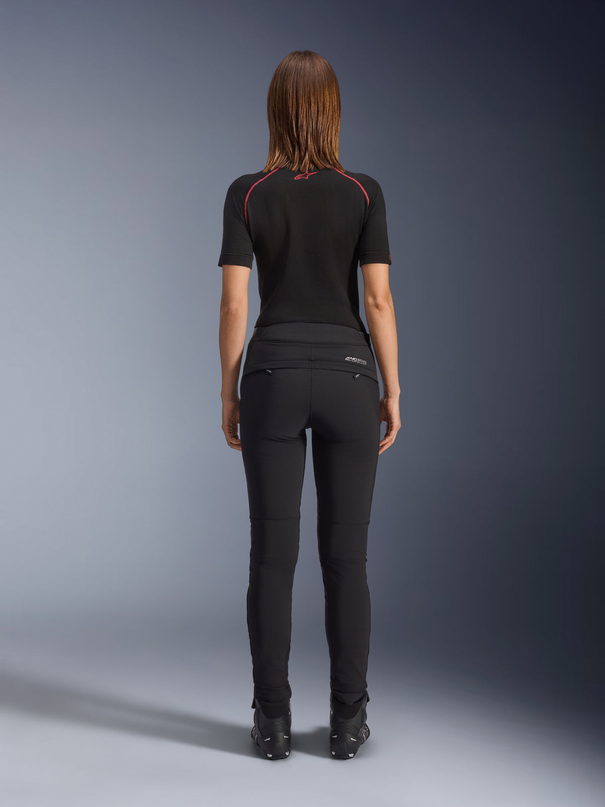 Alpinestars Alamosa Women Pants, nero, vista posteriore che mostra la struttura tessile slim-fit, tasche posteriori con cerniera e marchio MO.ST.EQ. riflettente sul carré posteriore, indossati con base layer nero a maniche corte e stivali da moto neri