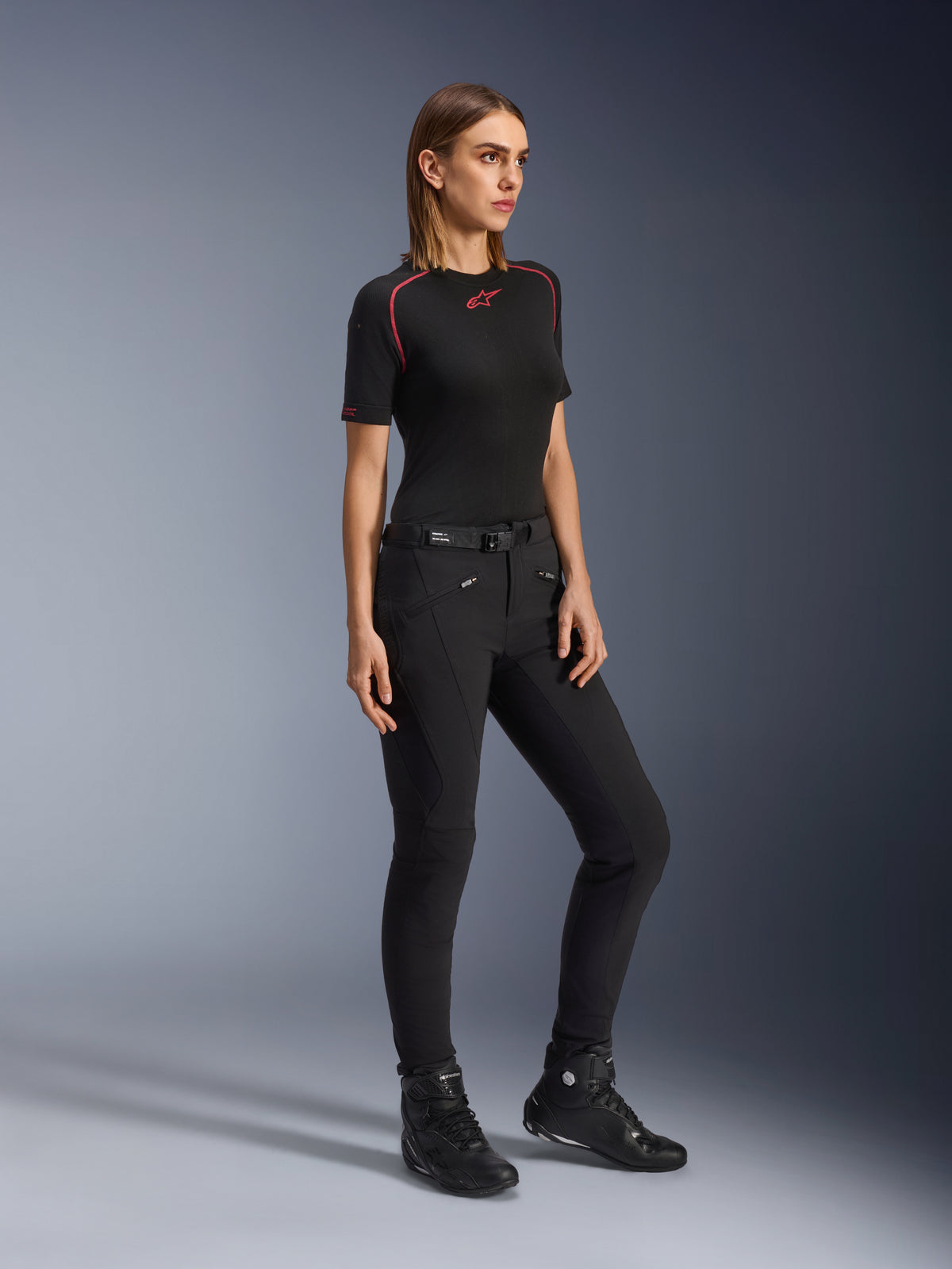 Pantaloni Alpinestars Alamosa Donna, pantaloni da moto slim-fit neri, indossati da una modella con un base layer nero e scarpe da moto, con tasche anteriori con zip e cintura con chiusura magnetica, stile streetwear urbano