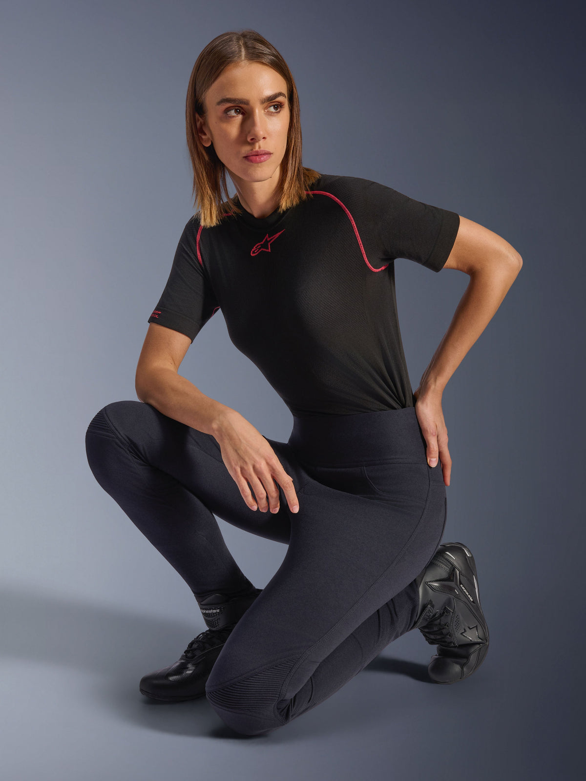 Leggings Alpinestars Stella Flex-Ast, neri, indossati da una donna inginocchiata in uno studio, abbinati a un top tecnico nero a maniche corte con profili rosa e stivali da moto neri, con vestibilità a vita alta e pannelli flessibili per la guida urbana.