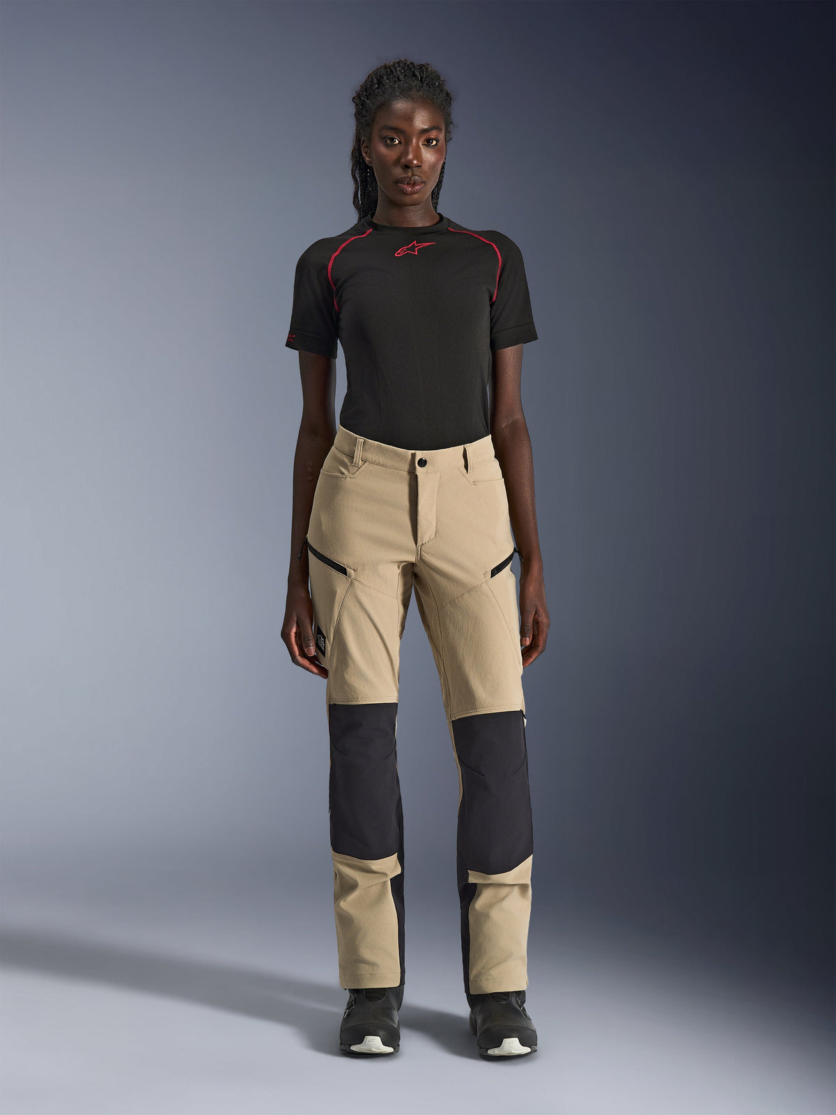 Pantaloni Alpinestars Stella Flex-AST Explorer da donna, pantaloni da moto, color sabbia con pannelli neri, indossati da una modella con maglia nera a maniche corte e stivali neri, dotati di pannelli rinforzati sulle ginocchia, tasche cargo con cerniera e tessuto ripstop traspirante per la guida adventure in climi caldi
