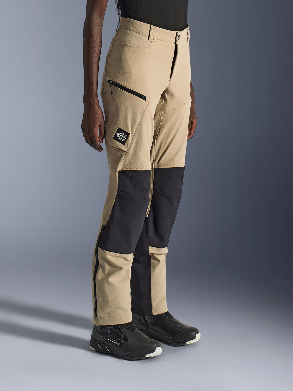 Donna che indossa i pantaloni Alpinestars Stella Flex-AST Explorer, pantaloni da moto adventure color sabbia e nero con pannelli ripstop neri sulle ginocchia, tasche cargo con cerniera, patch con logo 'ALL TERRA' e cerniere sulla parte inferiore della gamba.