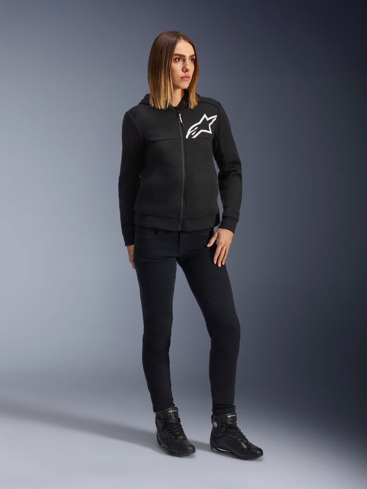 Donna che indossa Alpinestars Daisy V3 Women's Riding Denim, pantaloni da moto slim-fit in Rinse Black, abbinati a una felpa nera con zip e stivali da moto, abbigliamento protettivo casual urbano