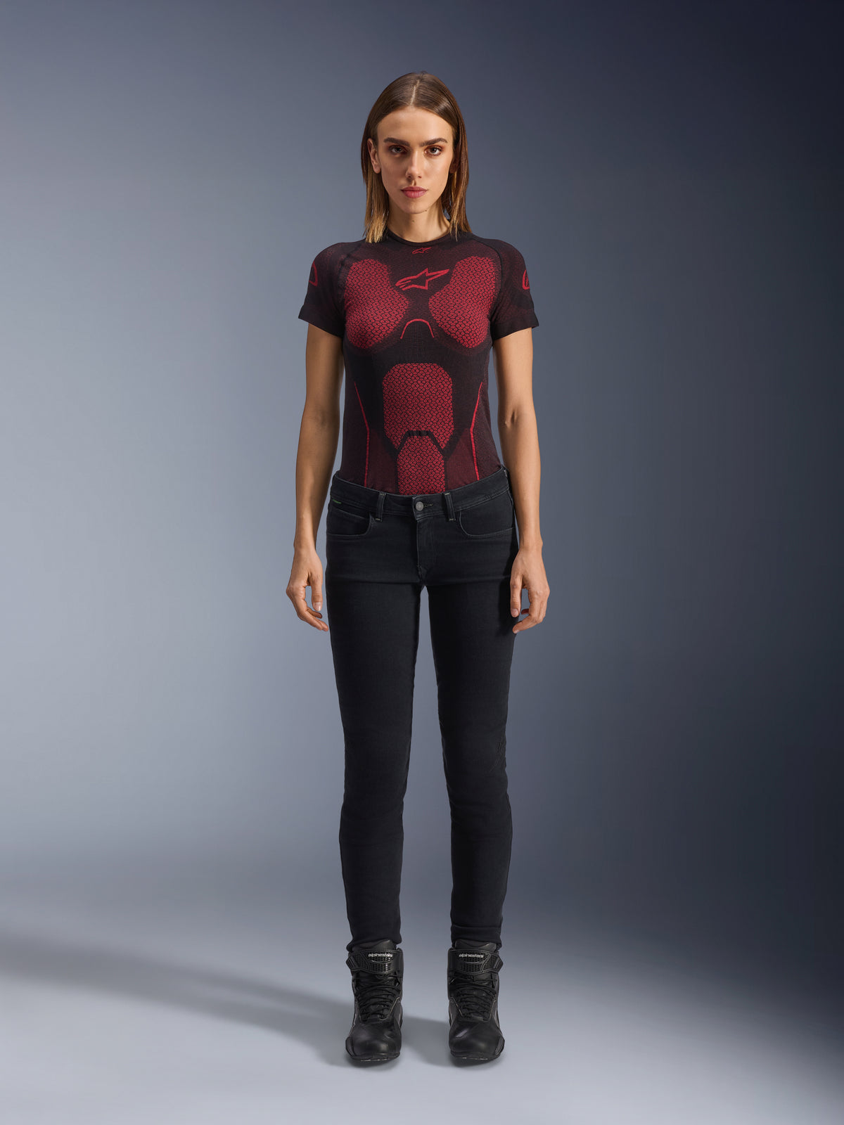 Alpinestars Daisy V3 Women's Riding Denim, pantaloni da moto slim-fit, nero, indossati da una modella con una t-shirt tecnica a motivi rossi e neri e stivali da moto neri, abbigliamento urban riding casual con silhouette femminile e design a vita alta.