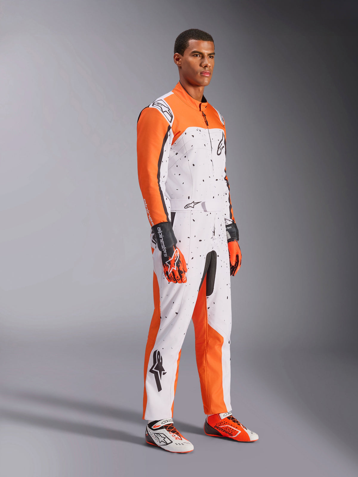 Alpinestars KMX-9 V3 Graphic 6 Karting Suit, bianco, arancione e nero, con motivo punteggiato e vivaci maniche arancioni, indossata da un modello con guanti e scarpe da corsa coordinati, abbigliamento professionale da motorsport a doppio strato.