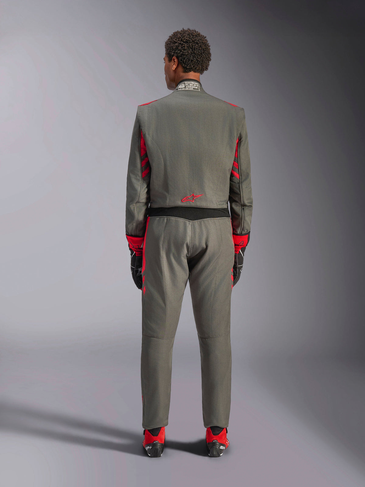 Vista posteriore di un modello che indossa la tuta Alpinestars GP Race V3 in Antracite Rosso, tuta professionale da auto racing con dettagli rossi su maniche e gambe, abbinata a stivali da corsa rossi e guanti neri, abbigliamento tecnico da motorsport in studio.