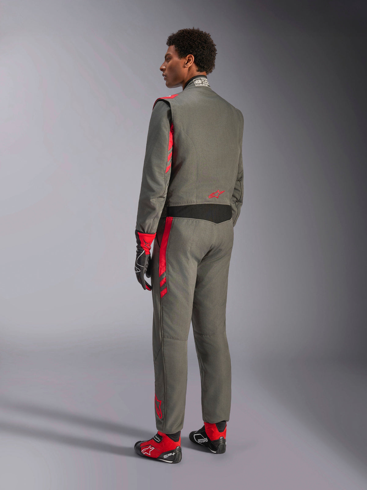Alpinestars GP Race V3 Suit FIA, tuta da corsa grigio antracite e rosso, vista posteriore del modello che indossa la tuta a due strati, accenti rossi su spalle e gambe, abbinata a guanti e stivali coordinati, equipaggiamento professionale per gare automobilistiche
