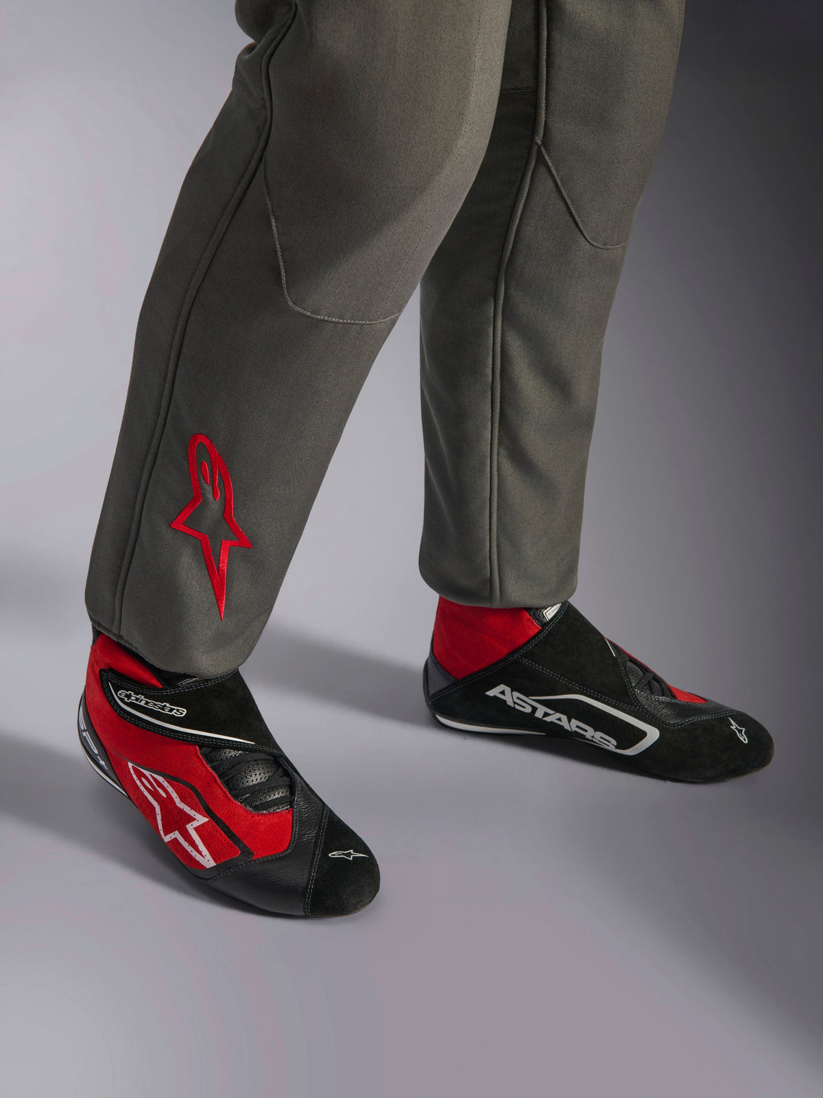 Alpinestars GP Race V3 Suit FIA, tuta da corsa Antracite Rosso, primo piano della parte inferiore delle gambe con logo a stella rosso ricamato, abbinata a stivali da corsa rossi e neri, abbigliamento automobilistico professionale progettato per la protezione e il comfort del pilota