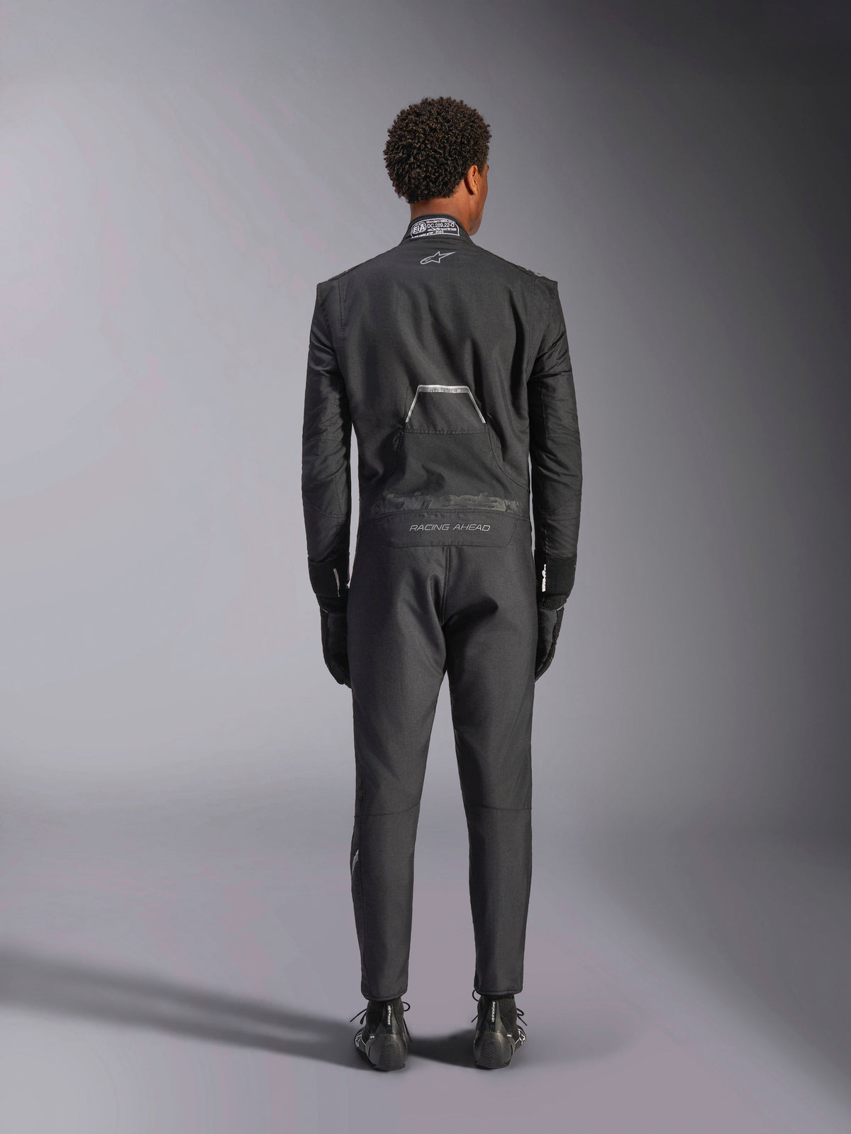 Tuta Alpinestars Hypertech V3, Black Dark Shadow, vista posteriore di una tuta intera professionale da auto racing, con etichetta di certificazione FIA sul colletto e marchi 'Alpinestars' e 'RACING AHEAD' sulla zona lombare, indossata con guanti e stivali da corsa neri coordinati.