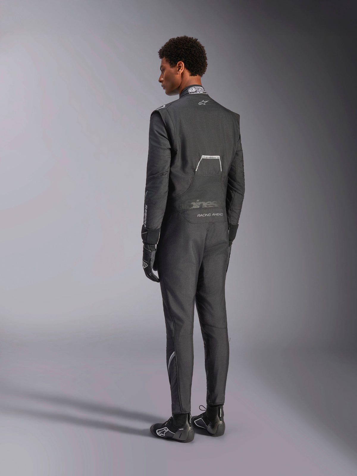Tuta Alpinestars Hypertech V3, Nero e Dark Shadow, vista posteriore di un modello maschile con tuta professionale da corsa auto, guanti e stivali neri coordinati, struttura leggera in Nomex, certificazione FIA sul colletto, vestibilità athletic-slim per il motorsport