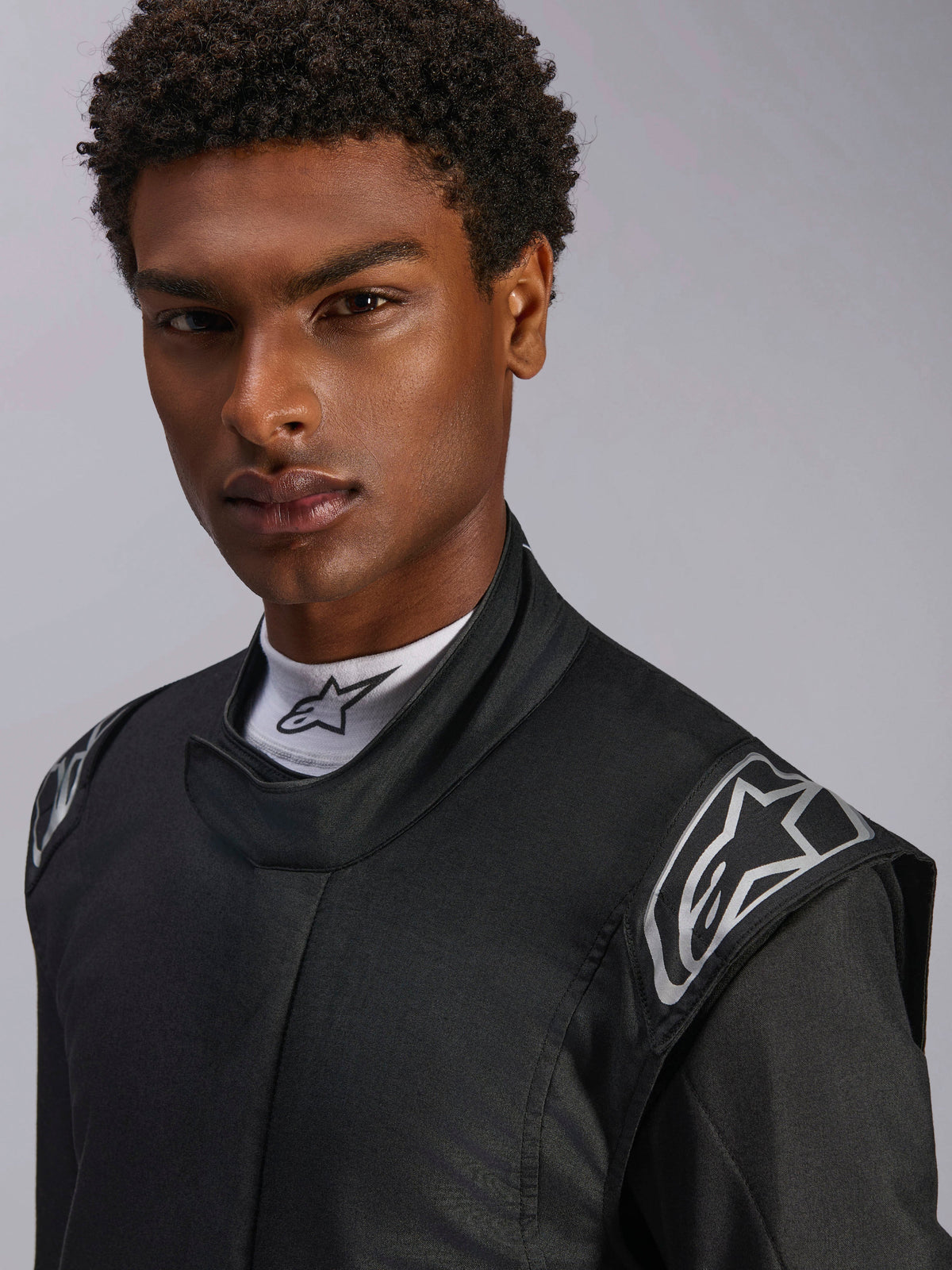 Tuta Alpinestars Hypertech V3, tuta da auto racing nero Dark Shadow, primo piano su modello, loghi argentati sulle spalle, colletto alto con maglia a lupetto bianca Alpinestars, abbigliamento protettivo professionale per motorsport