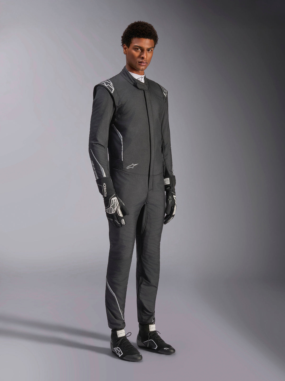 Tuta Alpinestars Hypertech V3, tuta da auto racing, nero e Dark Shadow, indossata da un modello con guanti neri e scarpe da corsa coordinati, vestibilità slim atletica, loghi Alpinestars e accenti grigio chiaro, abbigliamento protettivo professionale per motorsport