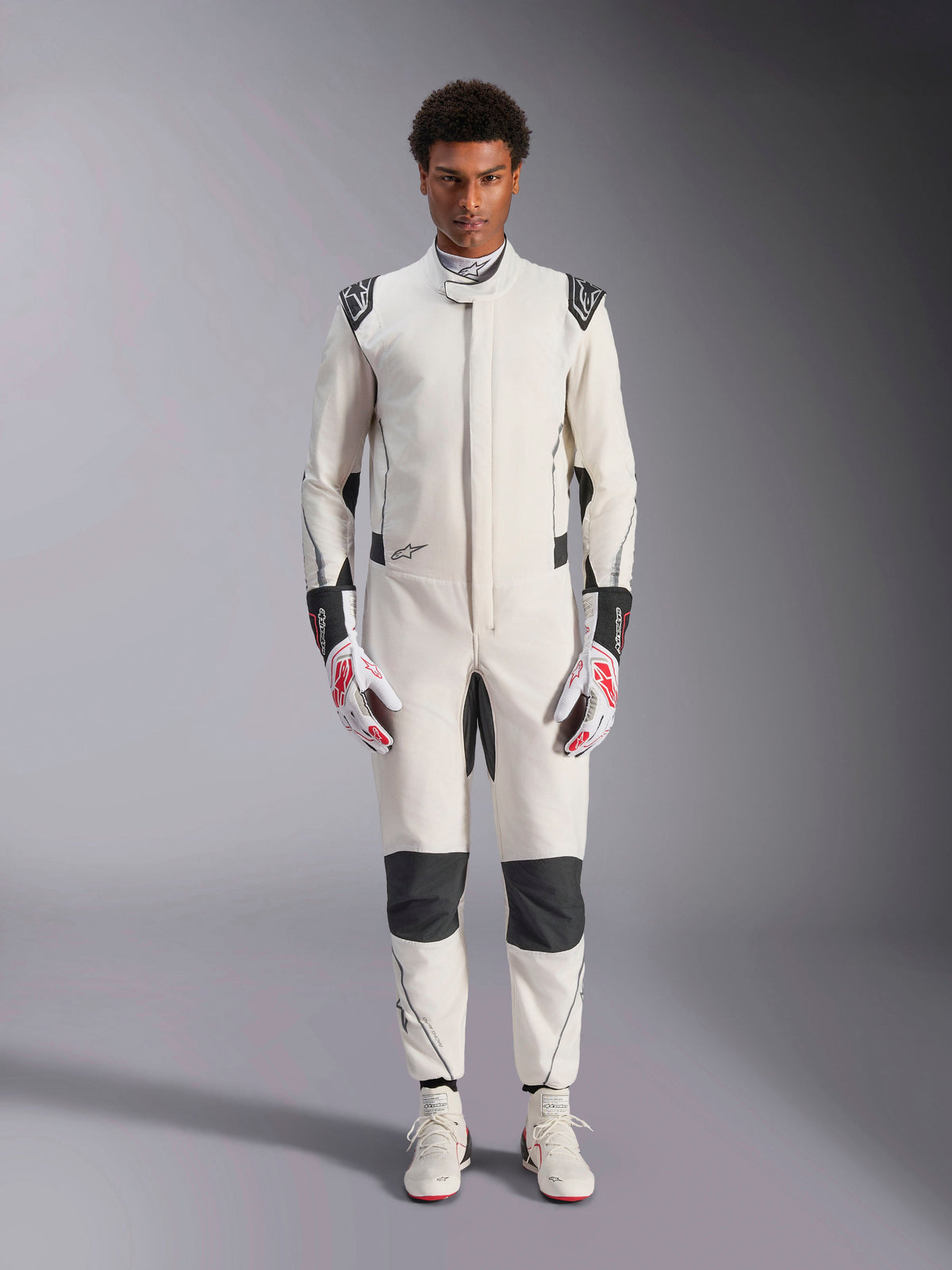 Tuta Alpinestars Hypertech V3, tuta professionale da auto racing, bianco, nero e argento, indossata da un modello con guanti rossi e bianchi e scarpe da racing bianche coordinate, caratterizzata da pannelli neri a contrasto e loghi Alpinestars, design con vestibilità atletica slim fit