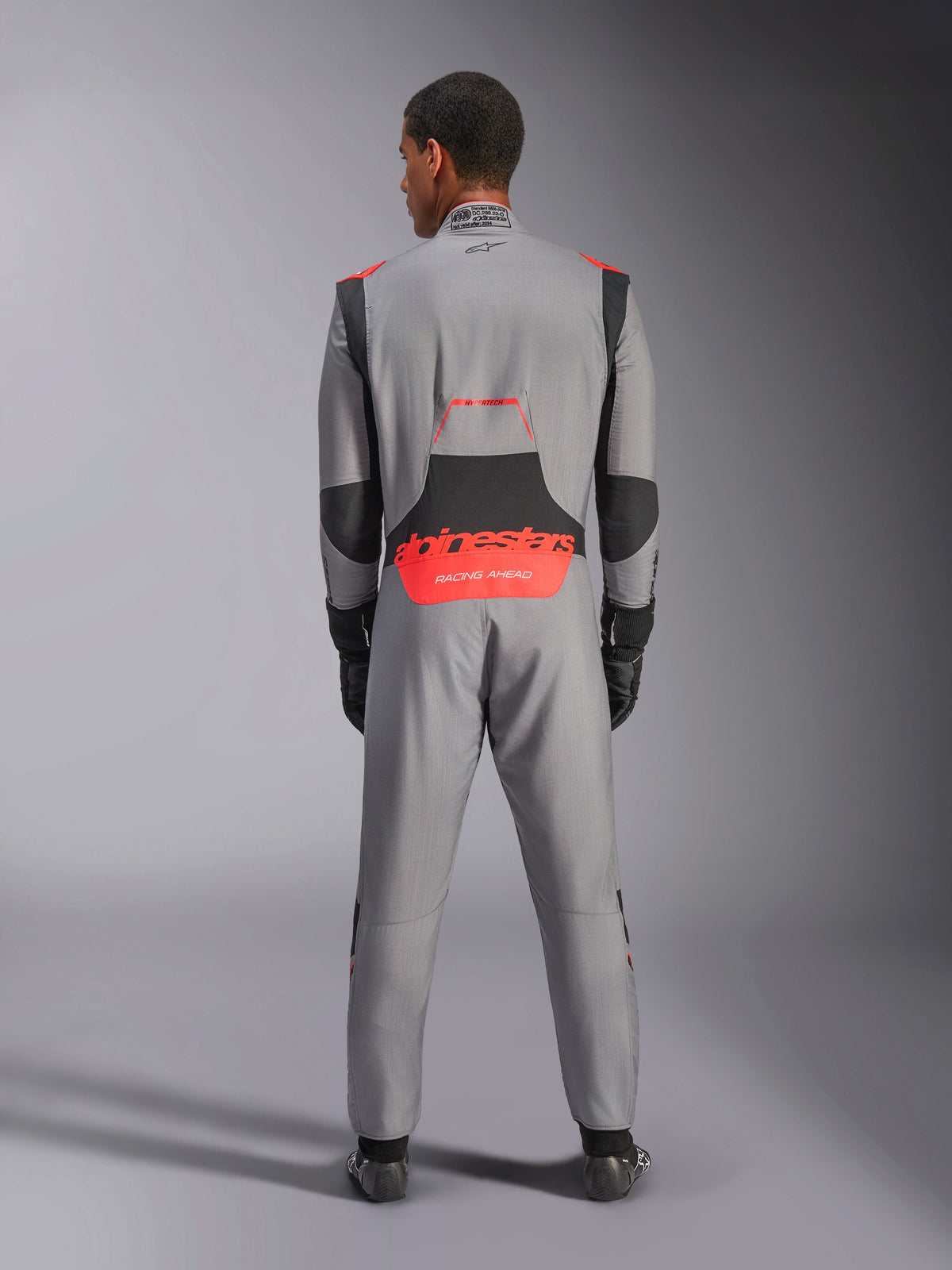 Tuta Alpinestars Hypertech V3, tuta da auto professionale, grigio medio con accenti nero e rosso fluo, vista posteriore su un modello con logo Alpinestars rosso sulla zona lombare, struttura leggera in Nomex per prestazioni in pista