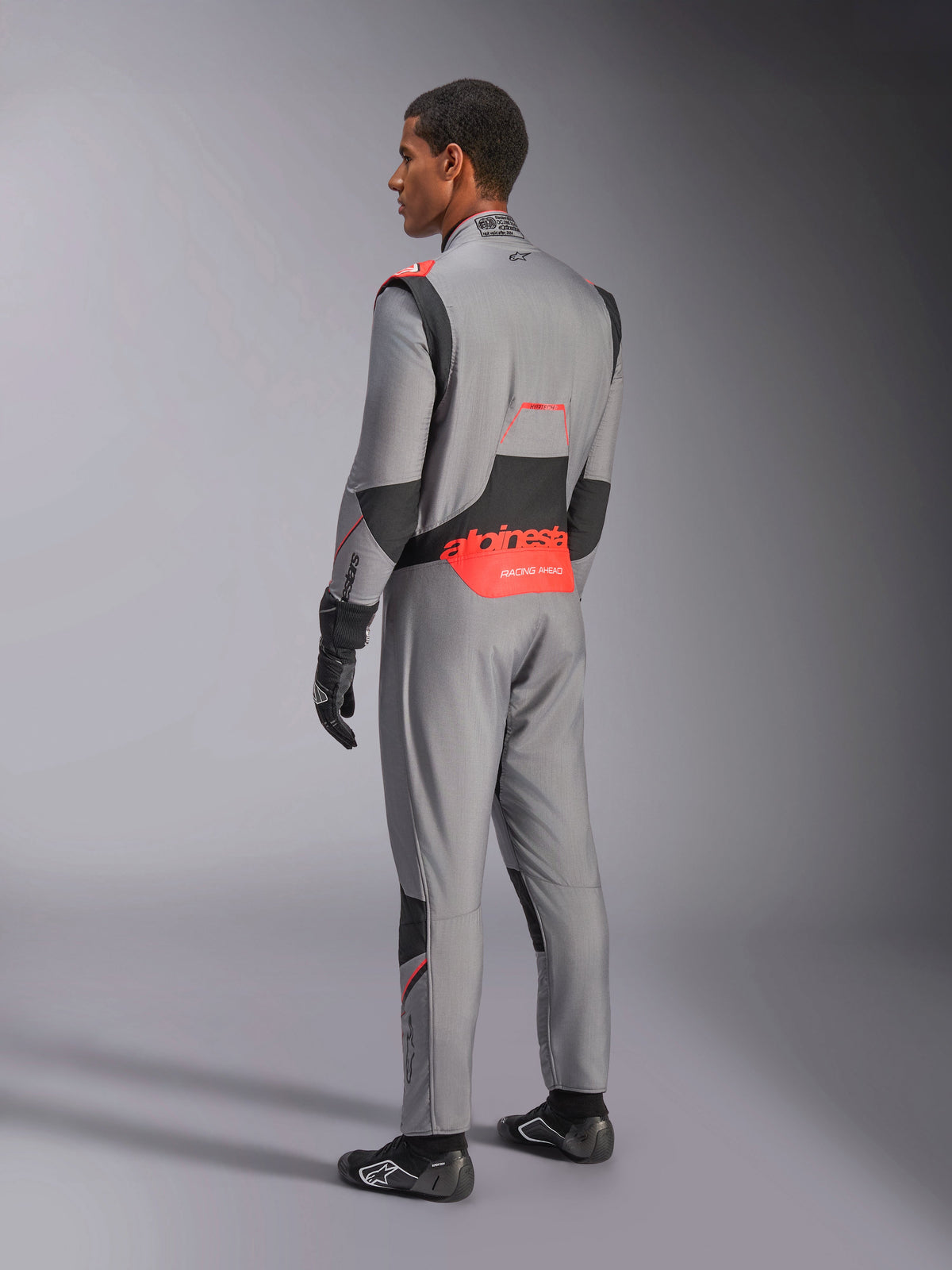 Tuta Alpinestars Hypertech V3 in grigio medio, nero e rosso fluo, vista posteriore di tre quarti, con logo Alpinestars rosso sulla parte bassa della schiena, pannelli elasticizzati neri e vestibilità atletica per competizioni automobilistiche professionali.