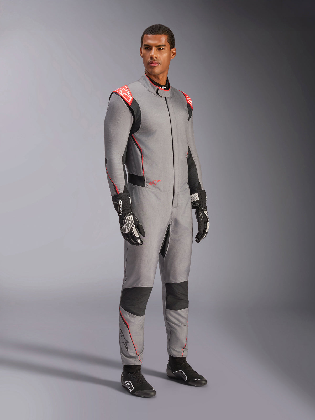 Modello che indossa la tuta Alpinestars Hypertech V3 in grigio medio con accenti neri e rosso fluo, tuta da corsa professionale abbinata a guanti e scarpe da racing neri, con struttura leggera in Nomex a due strati, dettagli rossi sulle spalle e vestibilità atletica