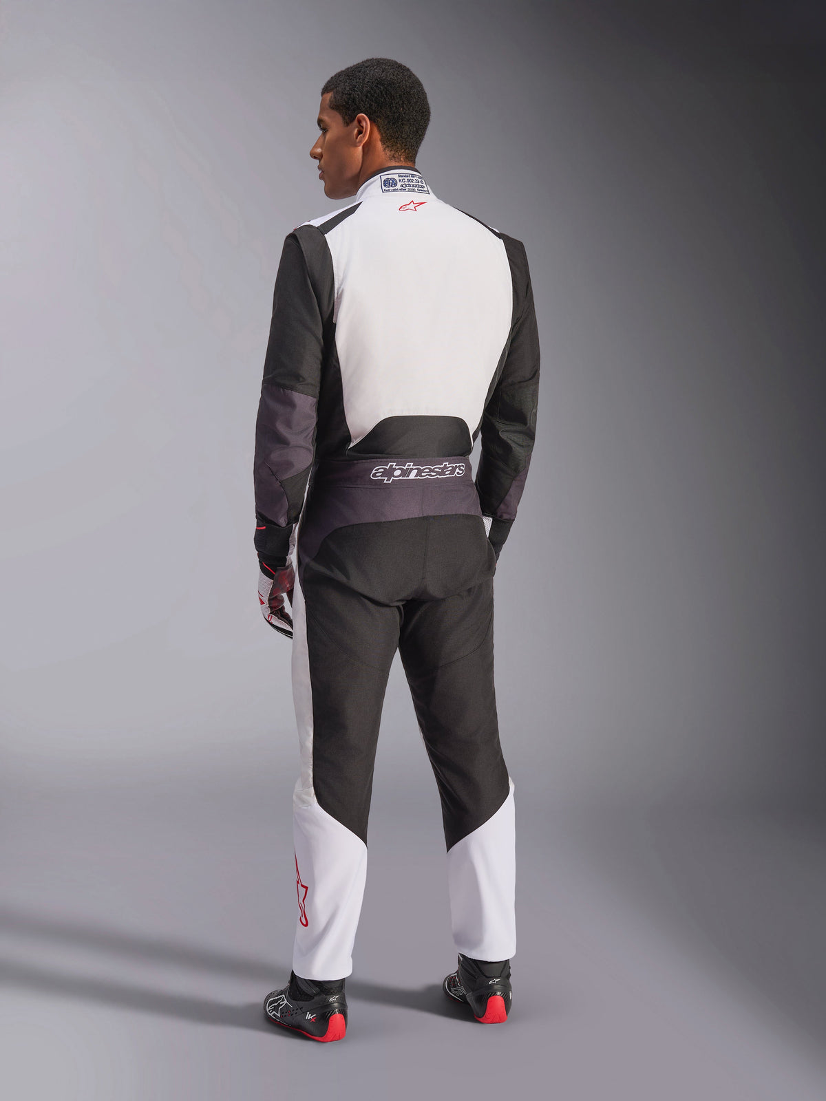 Alpinestars KMX-5 V3 Suit, tuta da karting, bianco, nero e rosso brillante, vista posteriore di un uomo che indossa una tuta da corsa intera con parte superiore della schiena bianca e pannelli inferiori neri, piccolo logo rosso sul colletto, indossata con stivali e guanti da corsa professionali