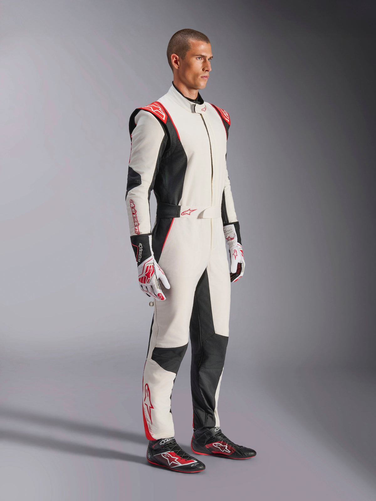 Pilota automobilistico che indossa la tuta Alpinestars GP Tech V4, bianco, nero e rosso, tuta intera da auto racing, guanti e scarpe da corsa coordinati, equipaggiamento professionale da motorsport con branding Alpinestars e design ergonomico
