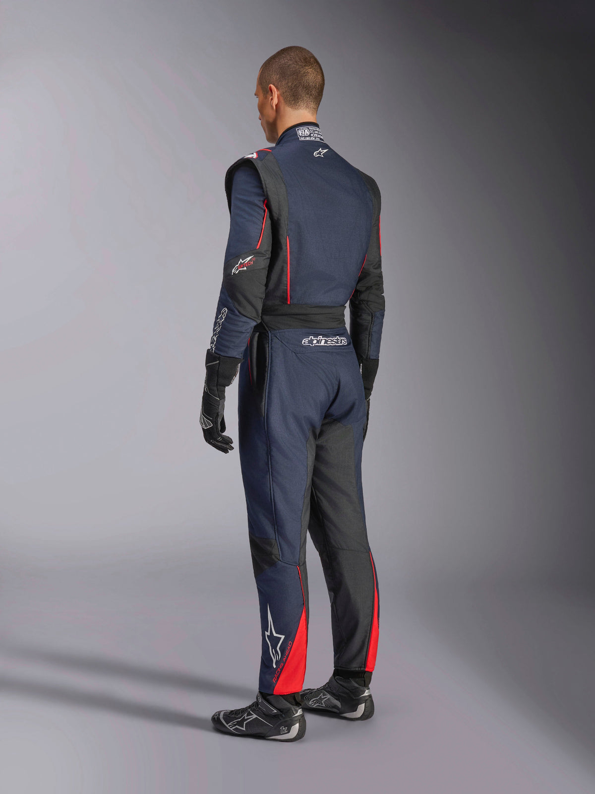 Tuta Alpinestars GP Tech V4, Blue Navy Nero Rosso, vista laterale posteriore di una tuta intera professionale da corsa, con accenti rossi e pannelli neri, indossata con guanti e scarpe da corsa neri, abbigliamento motorsport certificato FIA.